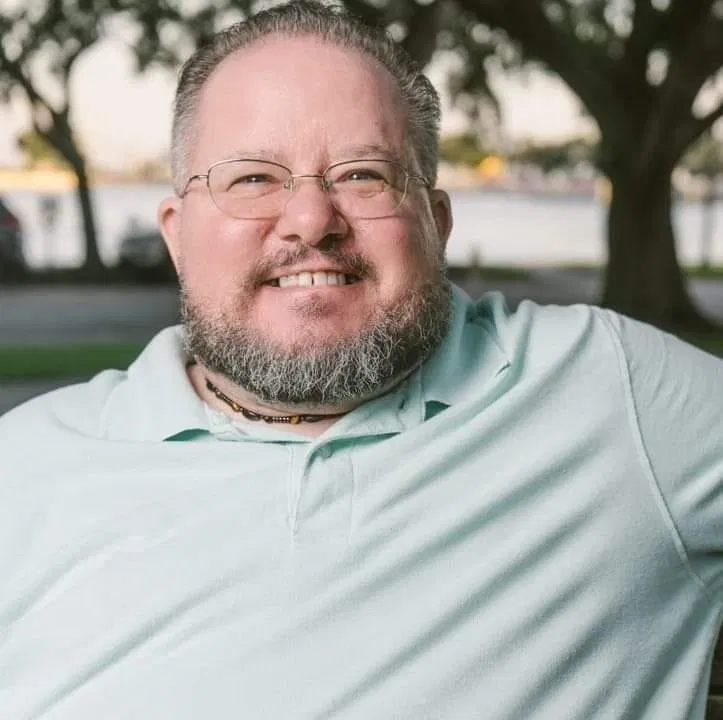 A smiling adult with a beard and glasses wears a light blue polo shirt, sitting outdoors with trees and water in the background. The image conveys warmth, approachability, and affirmation, reflecting our commitment to creating an inclusive and welcoming space for LGBTQ+ clients at Byrnes Counseling Group.