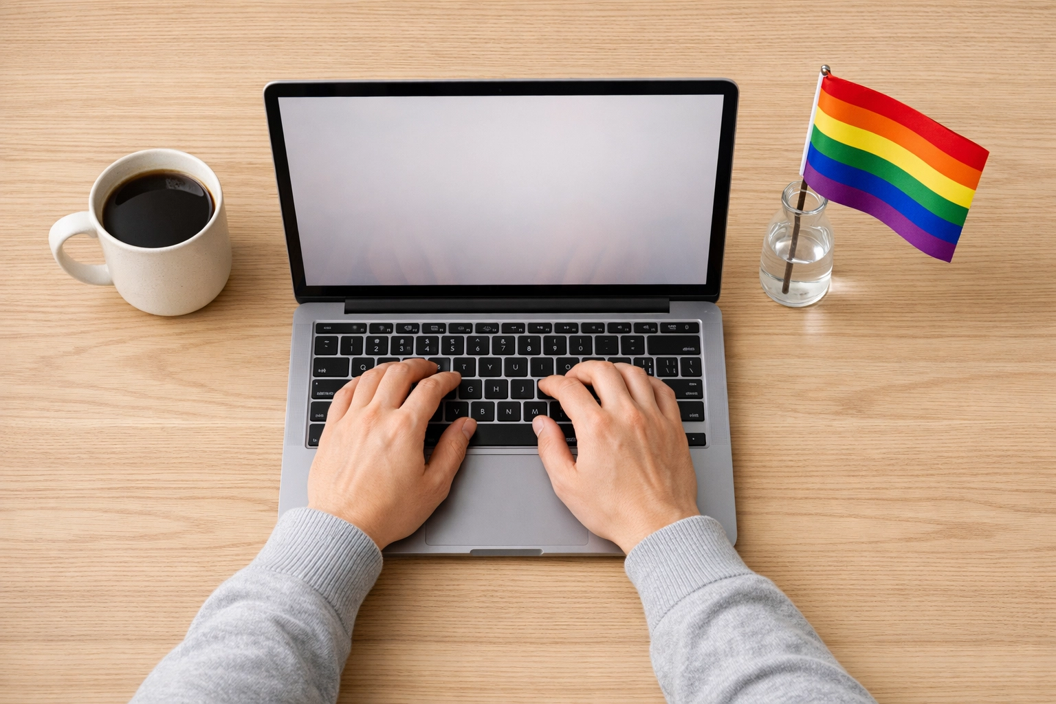 A queer author writing on a laptop next to a small pride flag, representing the future of LGBTQ+ independent publishing.