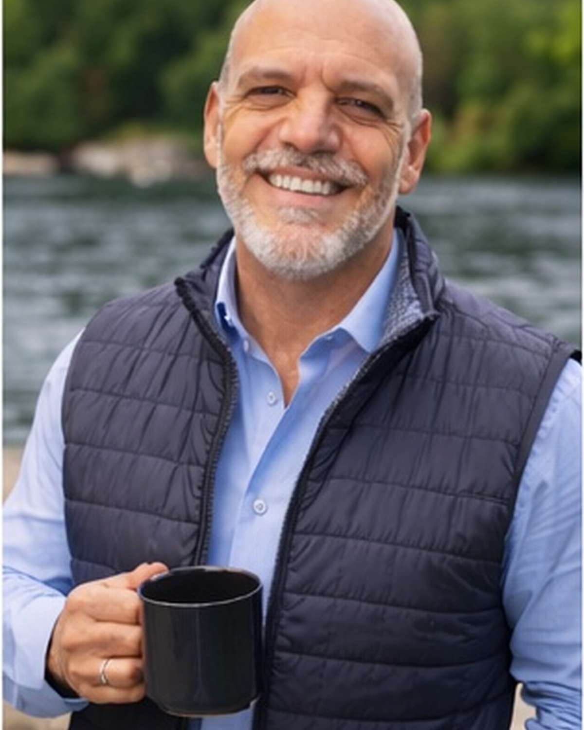 Smiling man in business casual attire holding coffee mug outdoors by waterfront