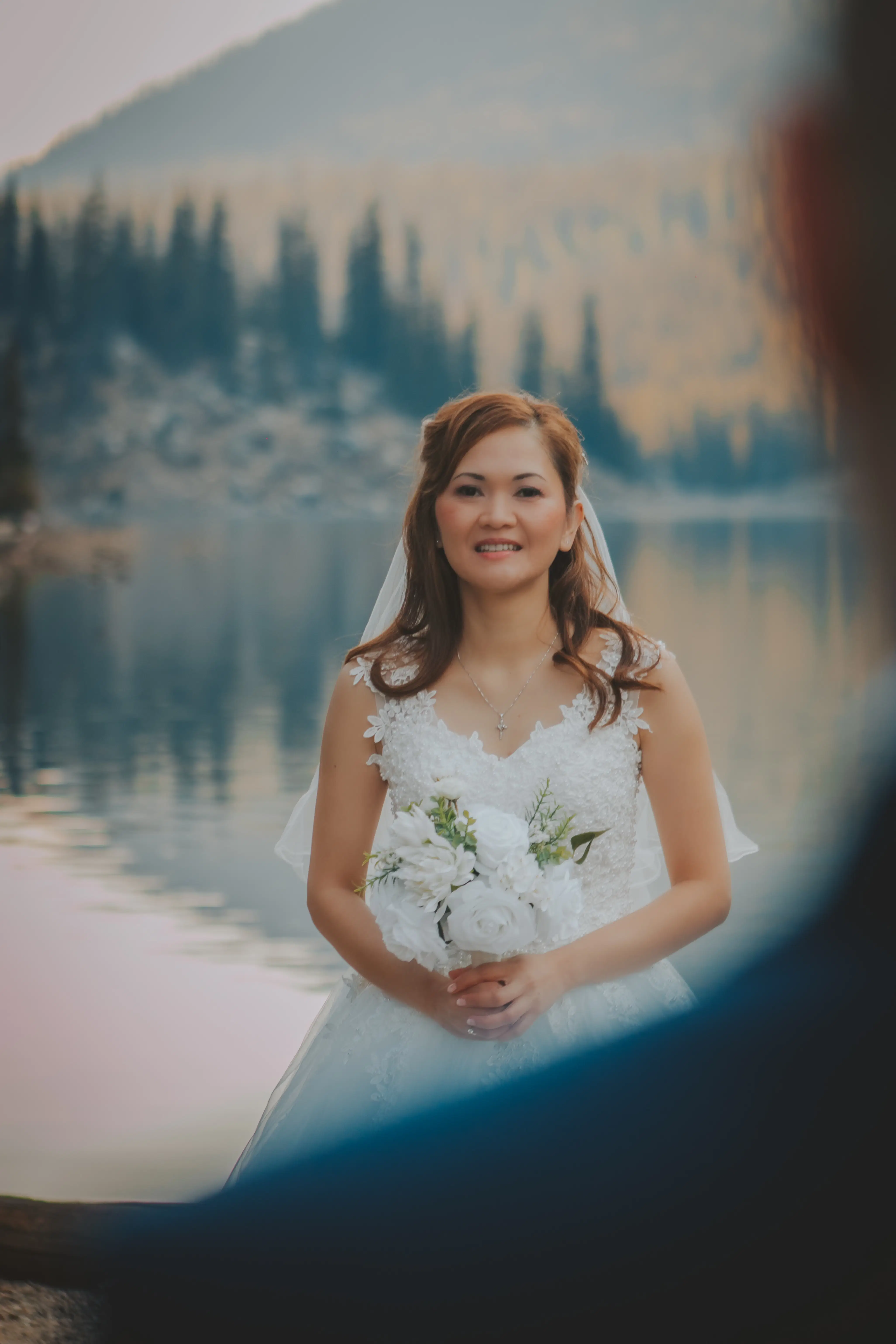 Bride in a lace wedding dress stands holding a bouquet of white flowers, with serene turquoise lake water and pine forest mountains in the background.