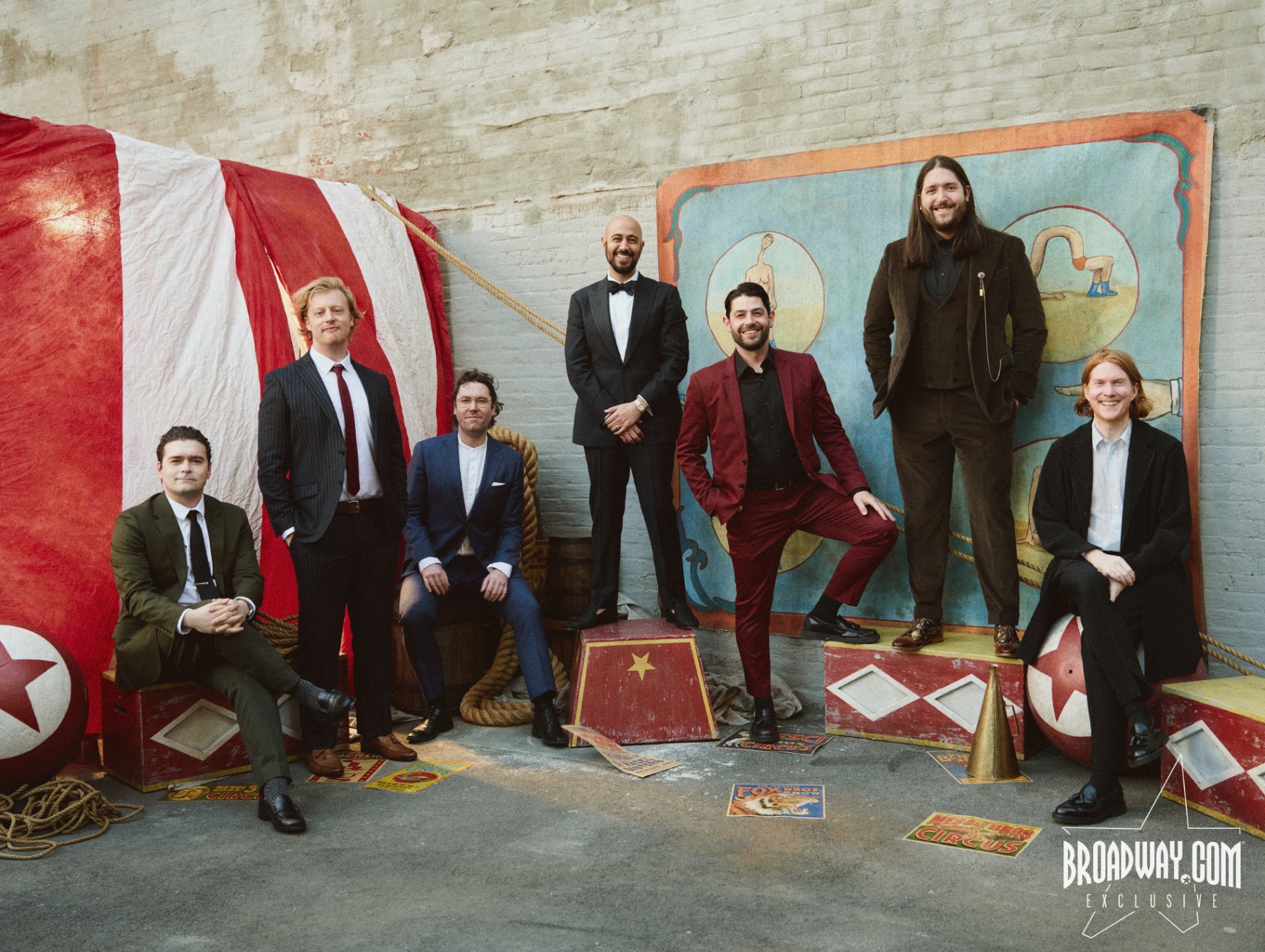 Seven men in formal and semi-formal attire pose in front of circus-themed props: a large striped tent, painted crates, ropes, a ball, and a backdrop with vintage illustrations. The setting suggests a creative ensemble cast participating in a theatrical or immersive performance incubated by The Orchard Project, capturing the project's spirit of artistic collaboration and inventive storytelling.