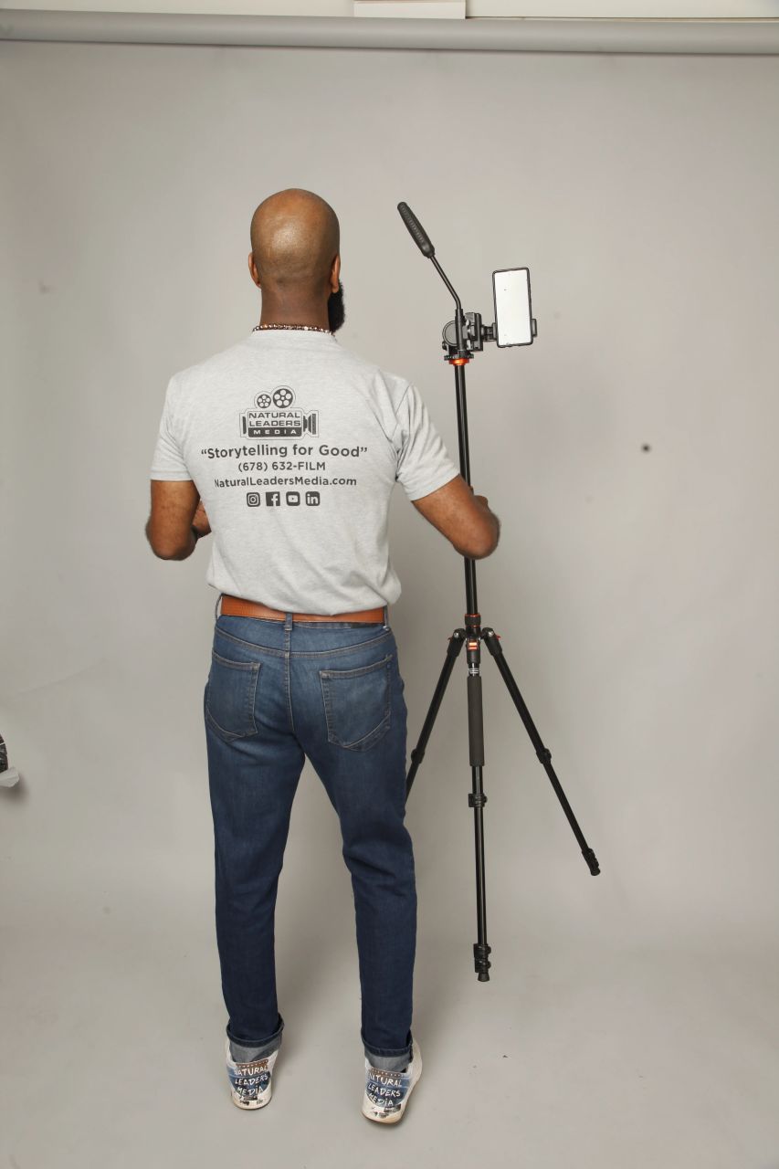 A Natural Leaders Media team member, wearing a branded 'Storytelling for Good' t-shirt, stands in a studio setting adjusting a smartphone mounted on a tripod: demonstrating professional mobile video production techniques as part of our educational workshops.