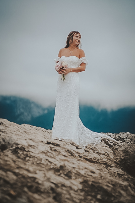 A bride in a white gown holding a bouquet and a groom in a dark suit share a joyful moment during their elopement ceremony on a forested lakeshore, with a pristine mountain lake and evergreen trees in the background, capturing the intimate, scenic experience offered by Tanan Banff Elopements.