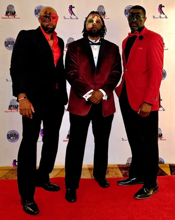 Three men in formal attire and decorative masquerade masks pose on a red carpet, highlighting the elegant and exclusive nature of a luxury group event.