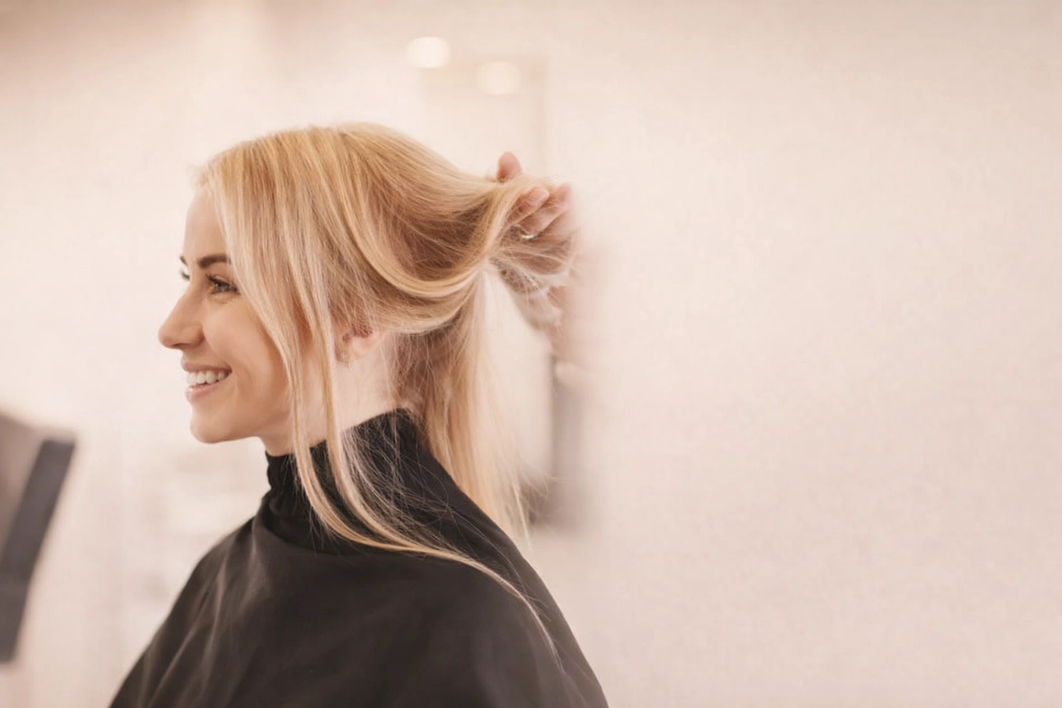 A smiling client with blonde hair sits in a cape at a modern salon while a stylist reviews her hair.