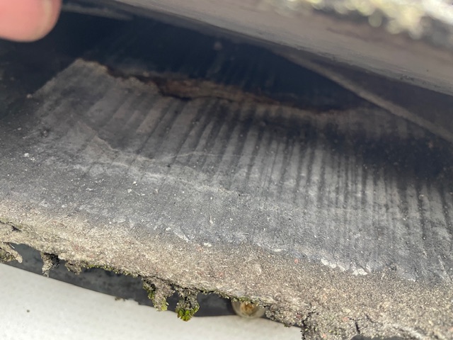Lifted asphalt shingle exposing asbestos tile underneath (moss/weathering visible)