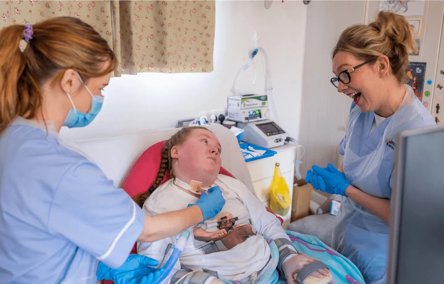 Two professional carers provide specialist medical care for a young woman with complex needs in a comfortable home setting.