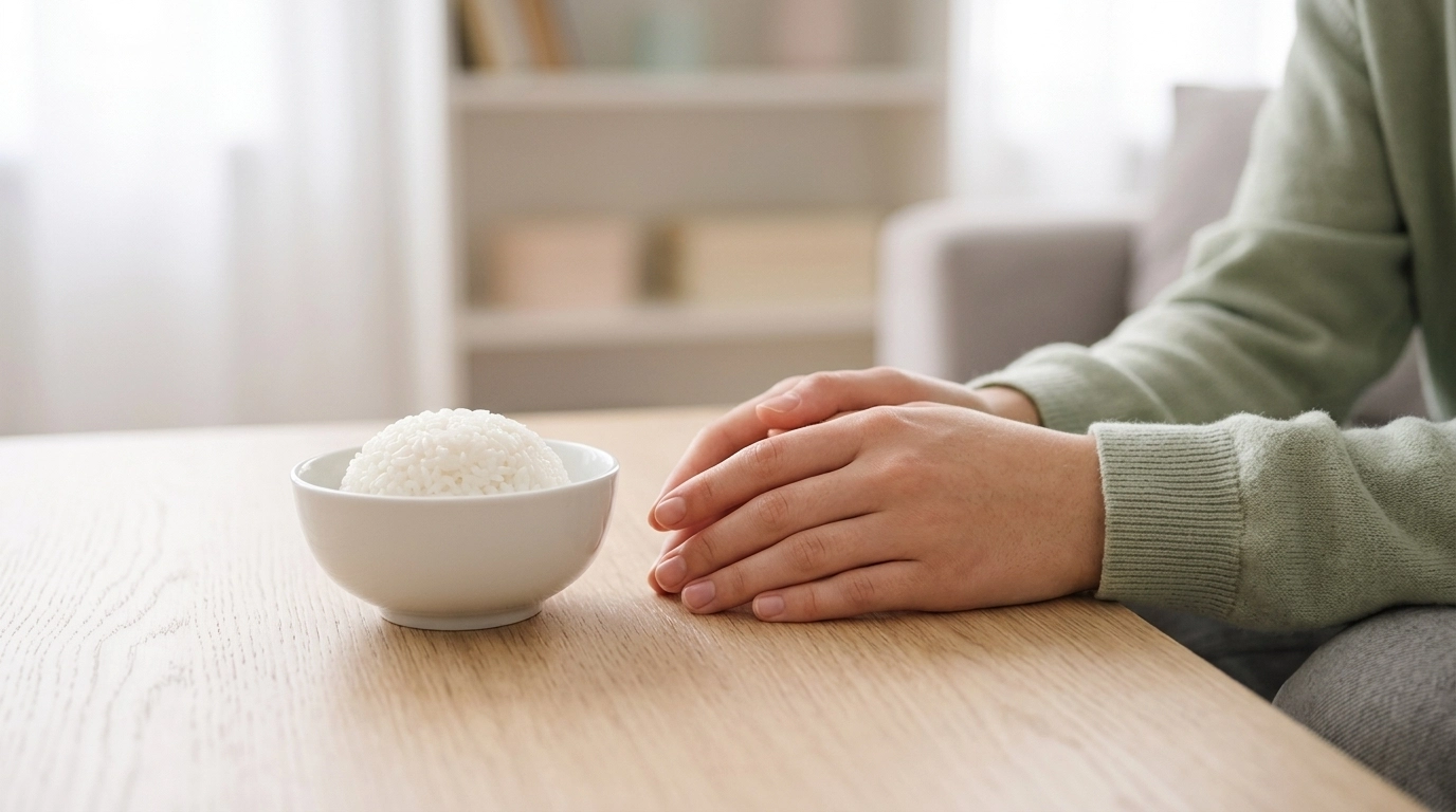 A minimalist photograph of a person's hands gently resting on a light-colored wooden table next to a small bowl of rice.