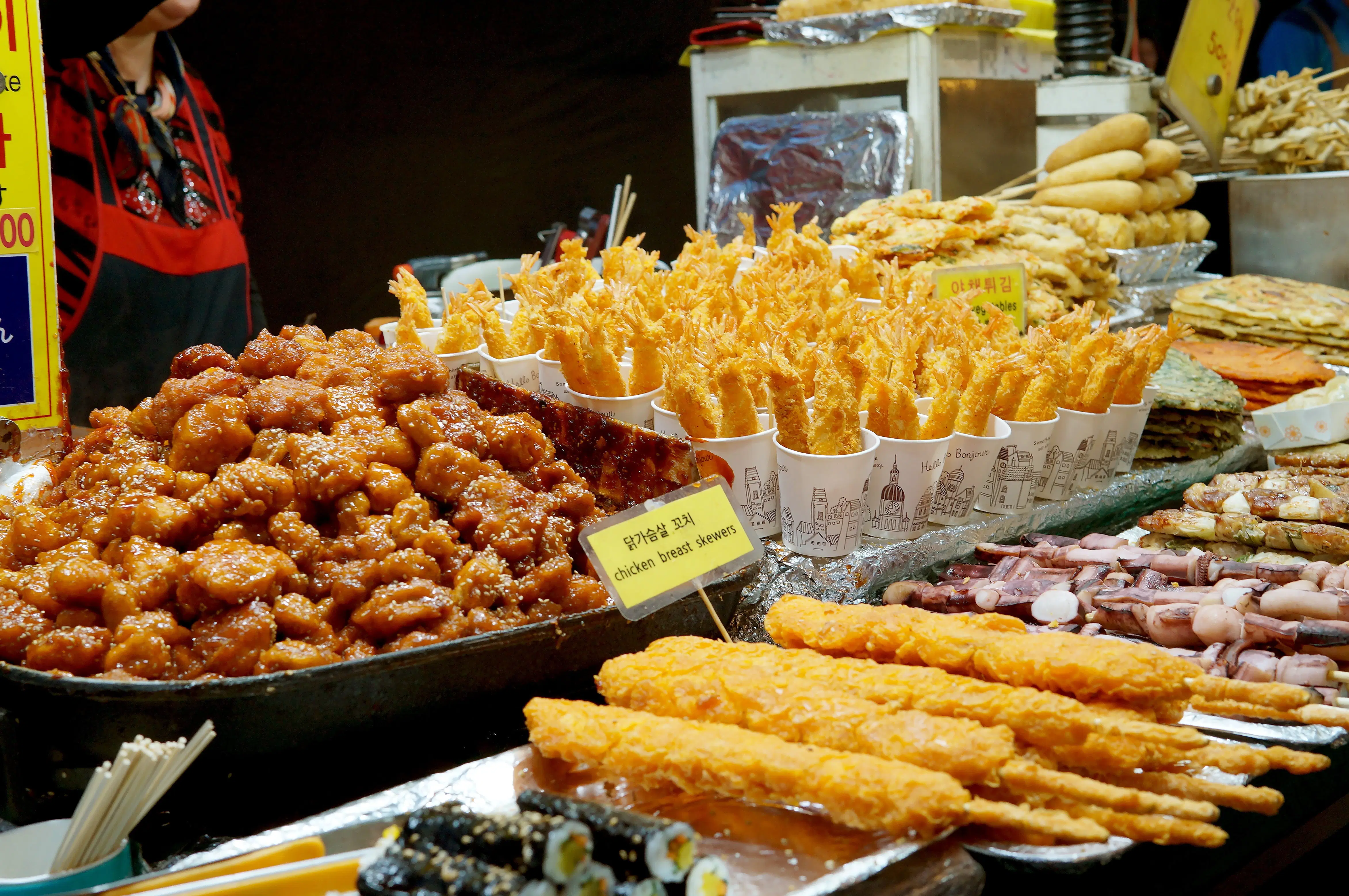 A vibrant festival food stall featuring crispy golden fried skewers, glazed chicken pieces, tempura prawns in takeout cups, kimbap rolls, and various grilled skewers from around the world.