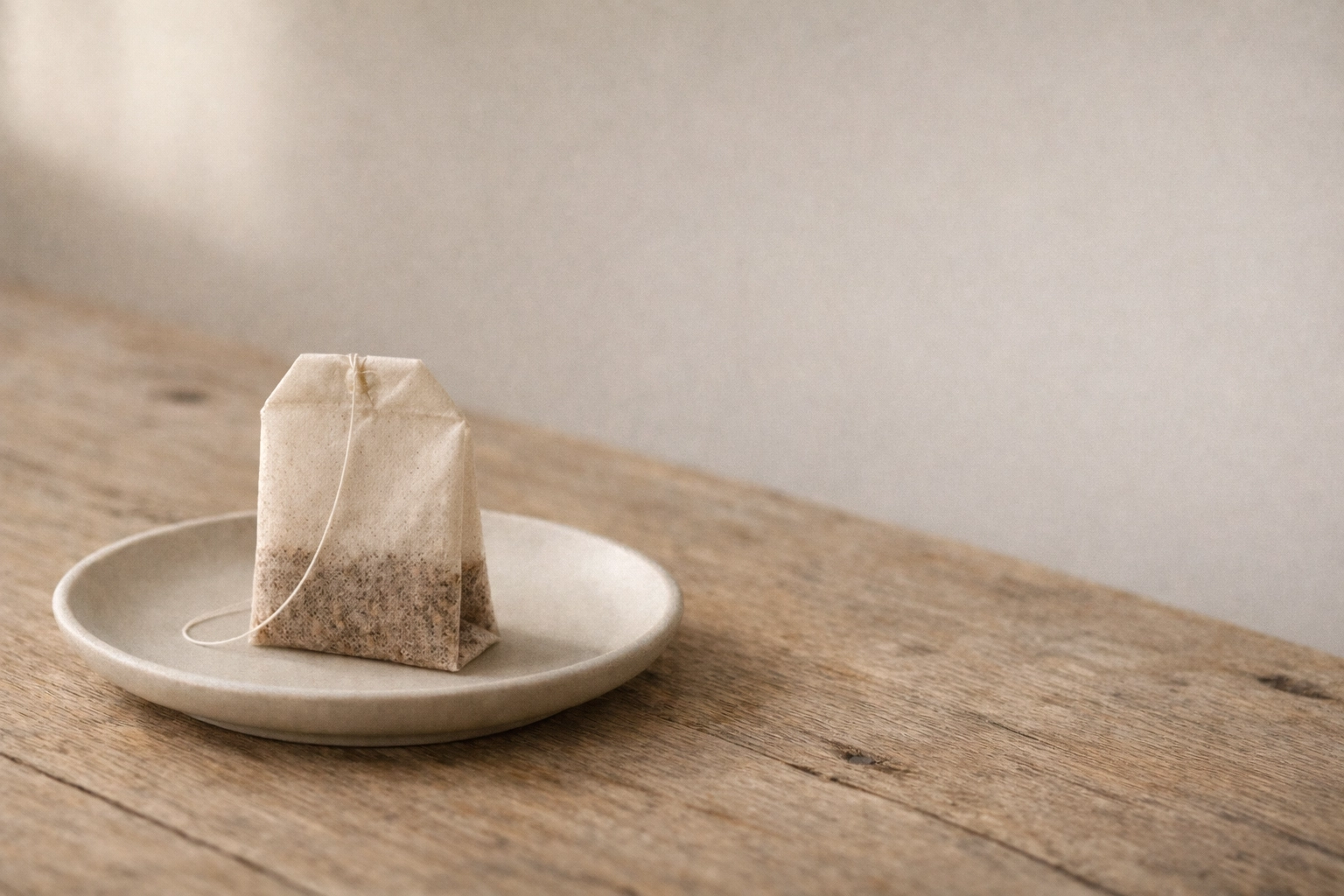 Close-up of a paper-fiber tea bag on a ceramic saucer in soft window light.