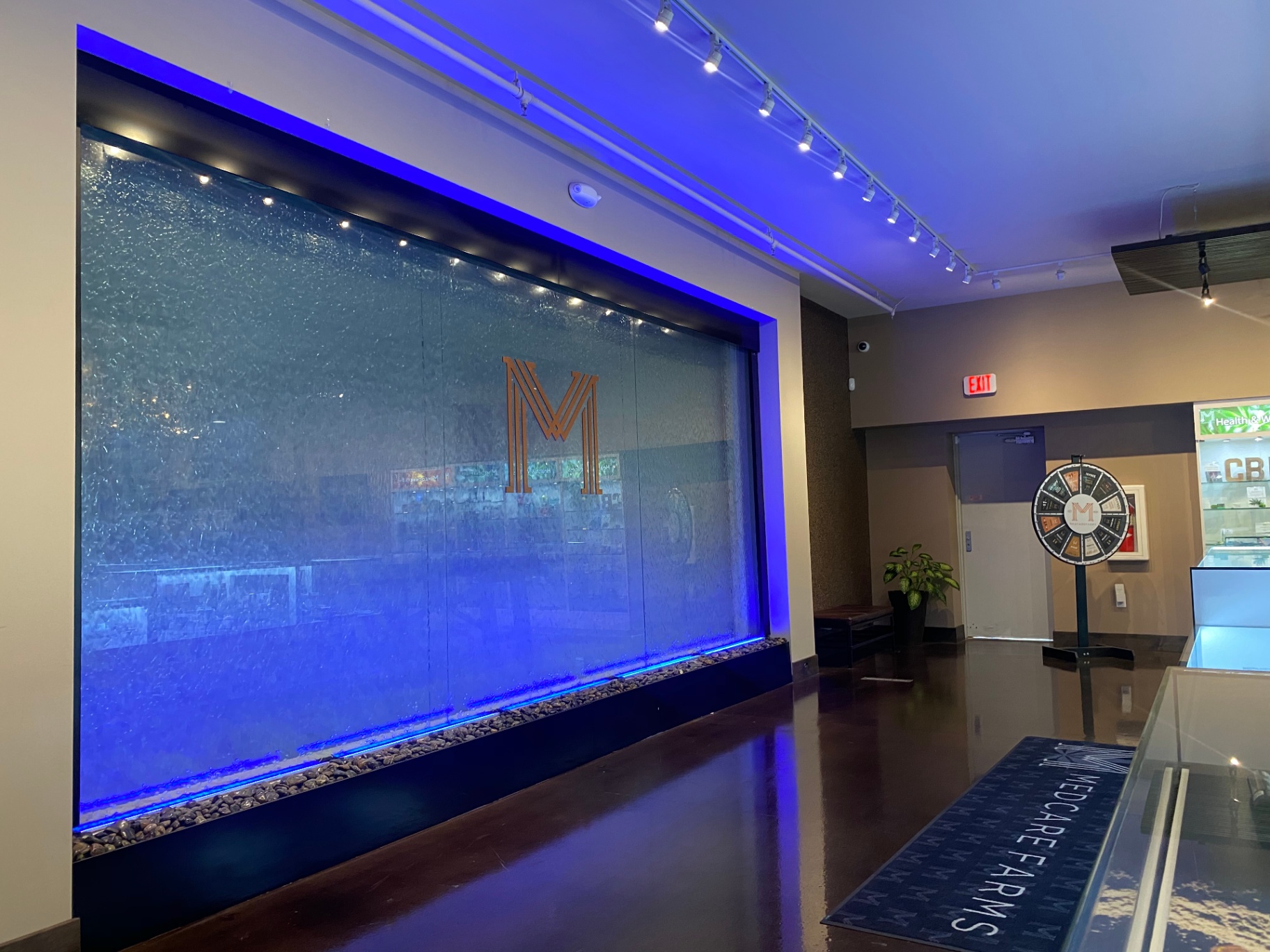 LL Waterfall Design Waterwall A custom-built, maintenance-free waterwall featuring L2O Liquid, illuminated with blue LED lighting for dramatic effect. The wall displays a large, central 'M' logo and is set in a modern commercial interior, making a striking visual statement at the entrance area.