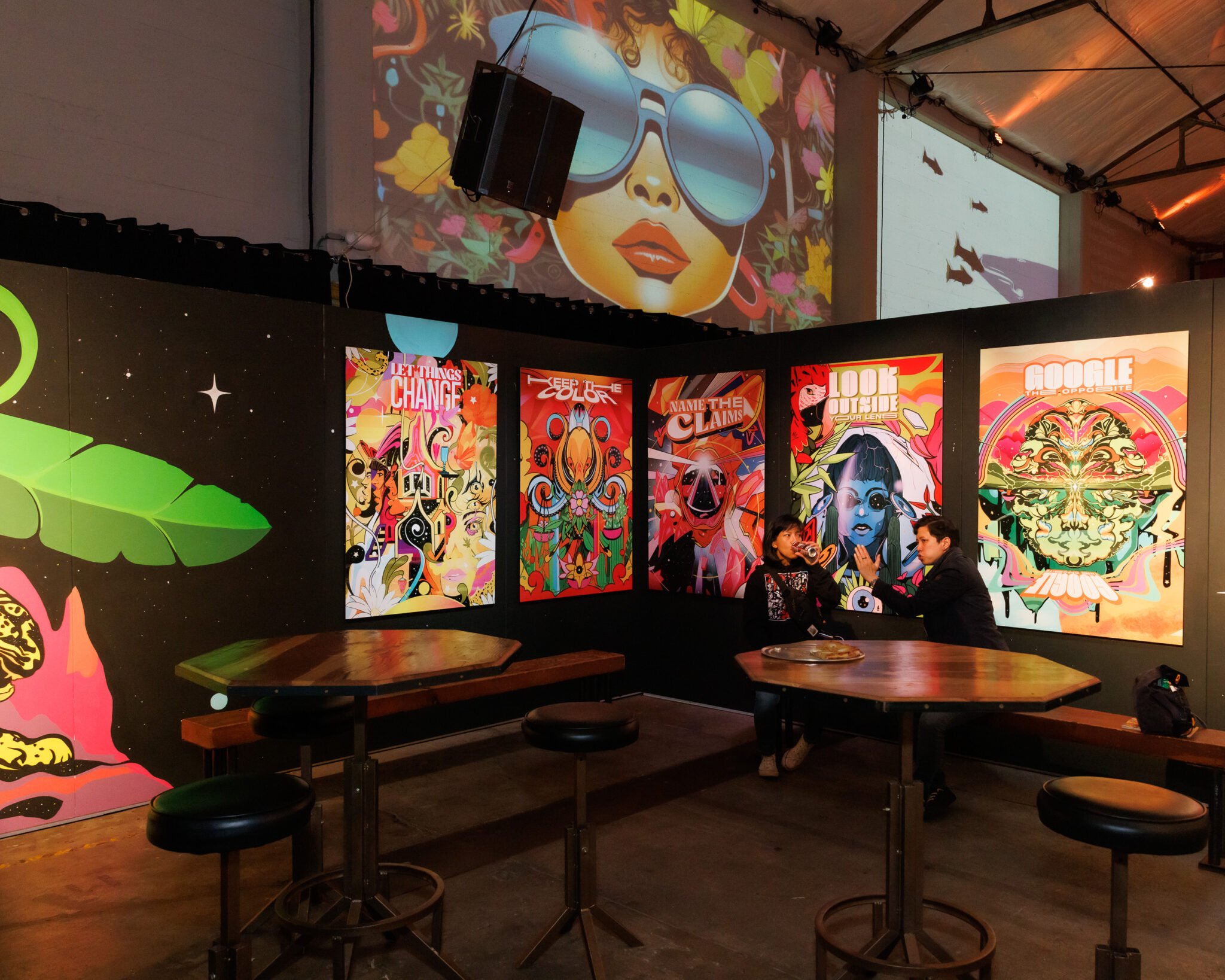 Industrial chic lounge area featuring geometric wood tables, black stools, bench seating, and bold contemporary art murals on black walls. The space includes a wall projection, colorful posters, and two people conversing, highlighting its versatility for casual networking or creative gatherings. Exposed beams and polished concrete floors underscore the venue's modern, adaptable atmosphere in SoMa.