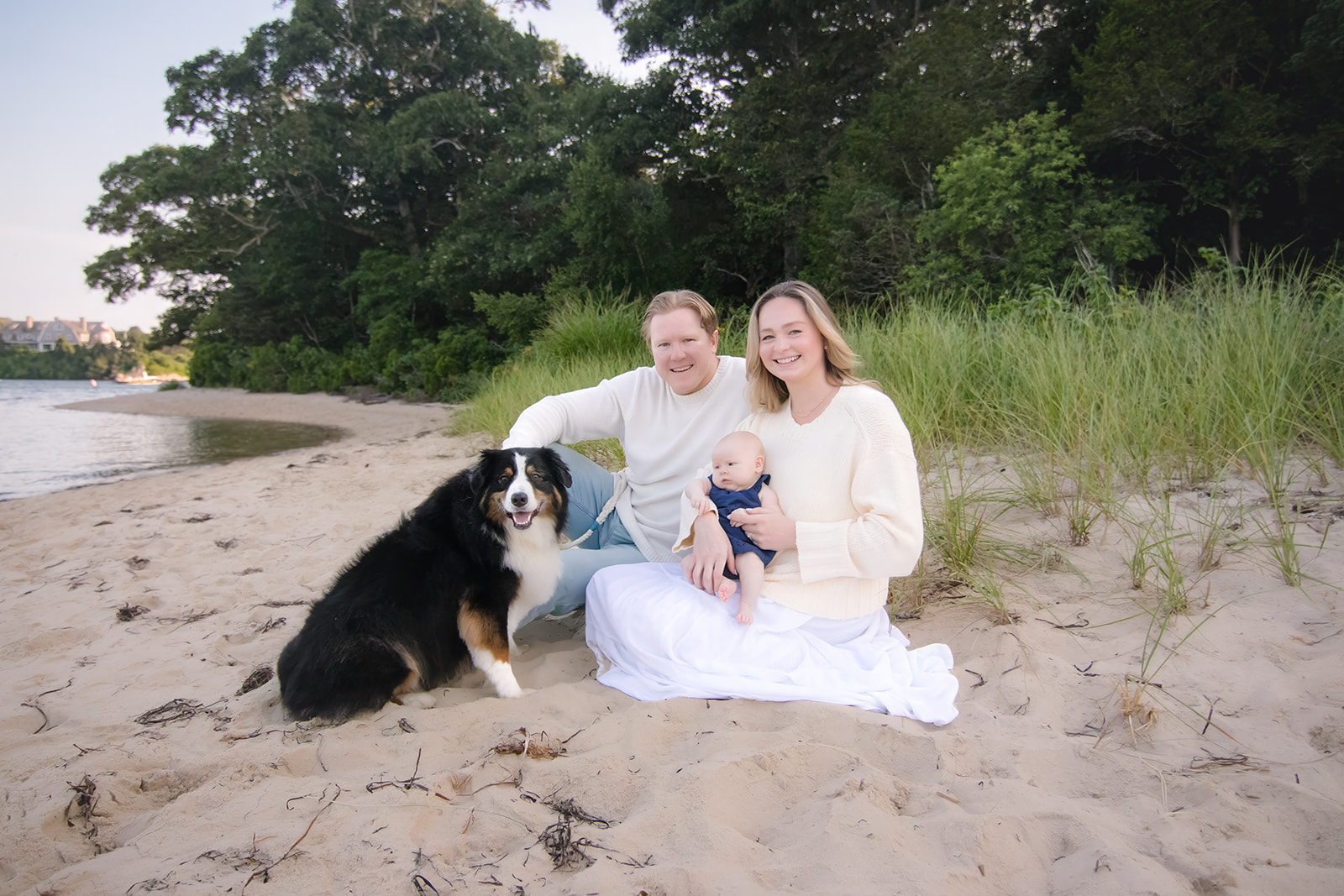 A young family sits together on a sandy Cape Cod beach with their baby and dog, surrounded by beach grass.