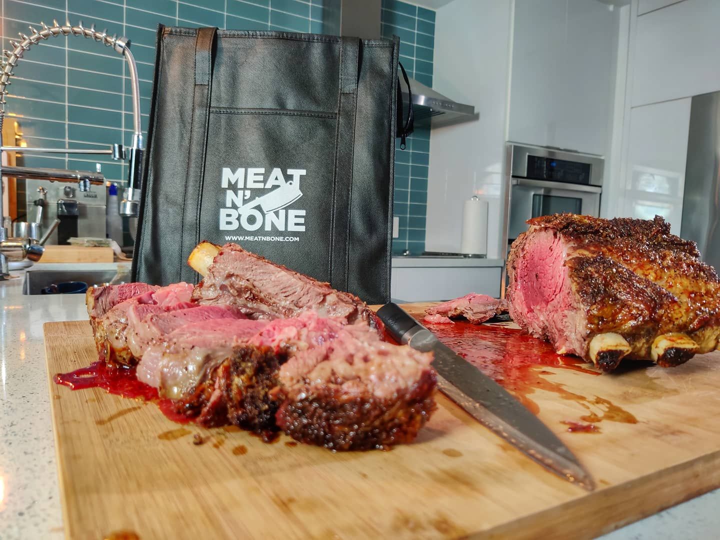 Sliced, juicy bone-in ribeye steak presented on a wooden cutting board, highlighting the marbling and doneness, with a Meat N' Bone branded bag in the background