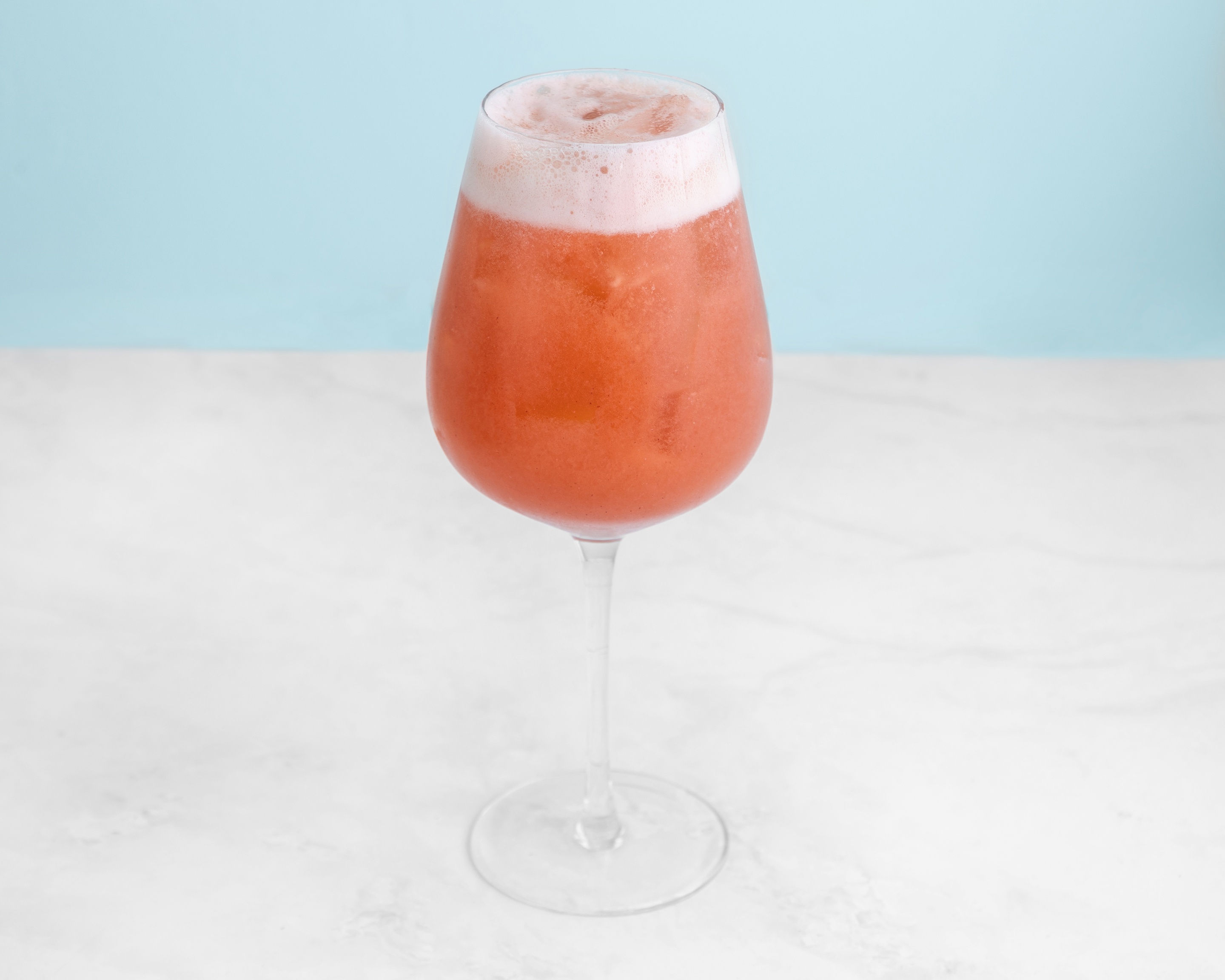 A stemmed wine glass filled with a foamy, ice-cold, pink-orange cocktail