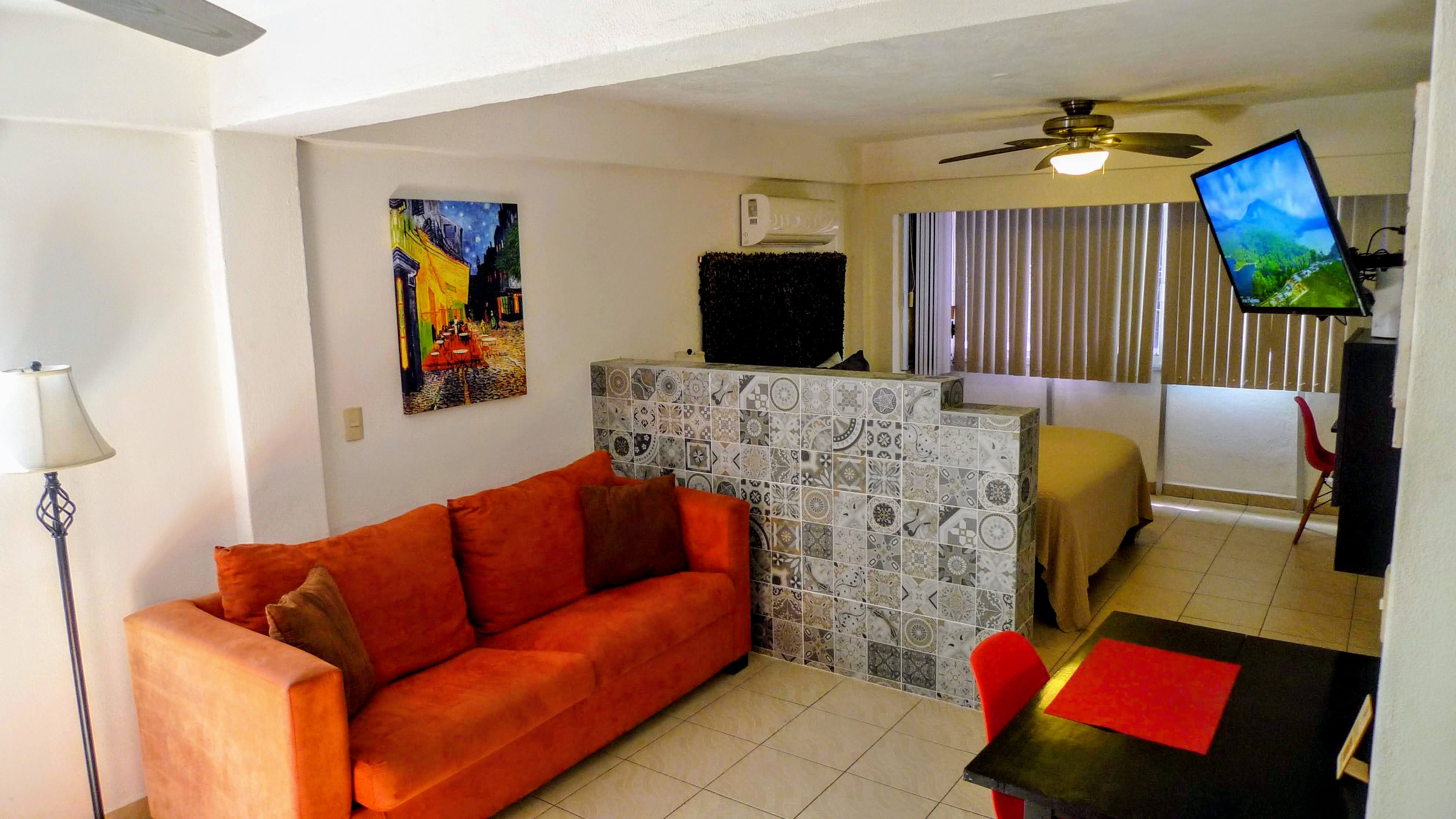 Modern Puerto Vallarta condo Open studio layout featuring a bright orange sofa, breakfast table, wall-mounted TV, decorative tile room divider, air conditioning, ceiling fan, and large windows with vertical blinds for natural light. Decor includes vibrant wall art and neutral tile flooring.
