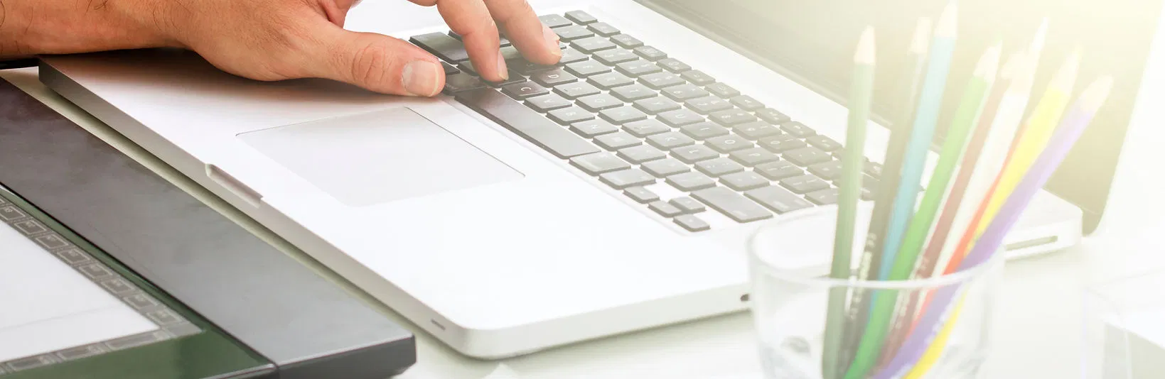 A hand uses a laptop on a tidy workspace next to a tablet and a cup of colored pencils, symbolizing remote work and IT support services.