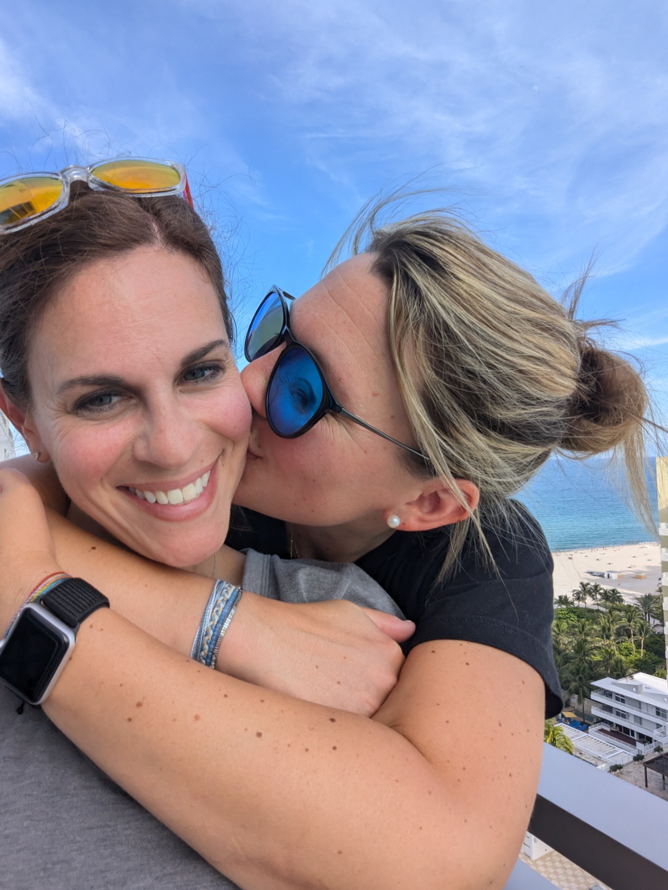 Two women share a joyful and affectionate embrace outside, one kissing the other on the cheek. Both are smiling and relaxed.