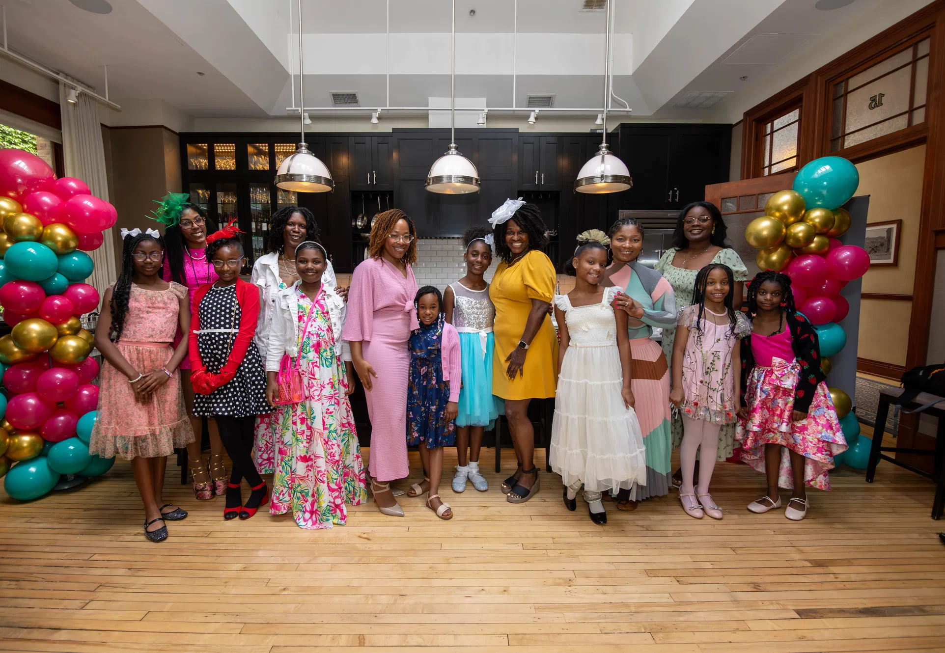 Diverse girls and mentors in formal dresses standing together in a festive, affirming atmosphere decorated with pink and gold balloons.