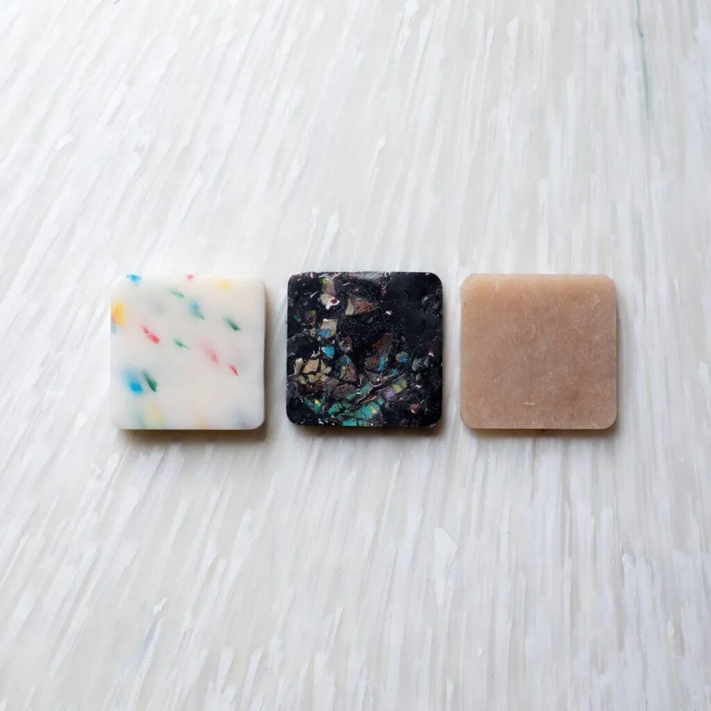 Three sample swatches of 100% recycled plastic panel material, displaying a white base with multicolored flecks, a black base with iridescent fragments, and a solid beige.