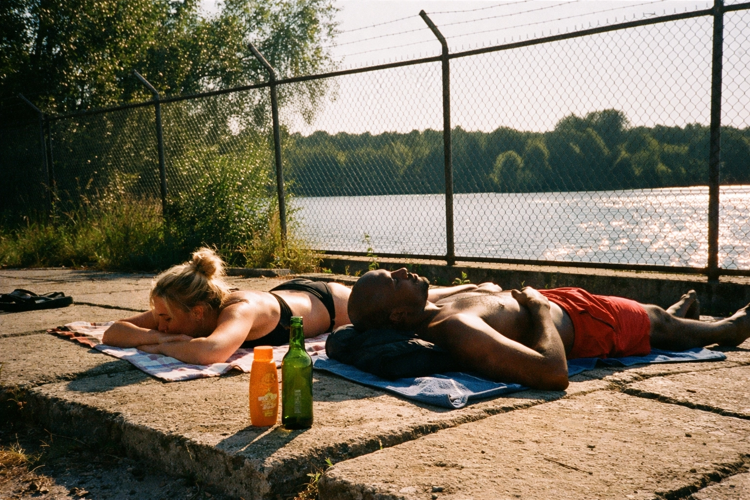 Flughafensee gritty DIY photo: tanning on concrete slabs by the water, industrial fence, beer bottle and cheap sunscreen.