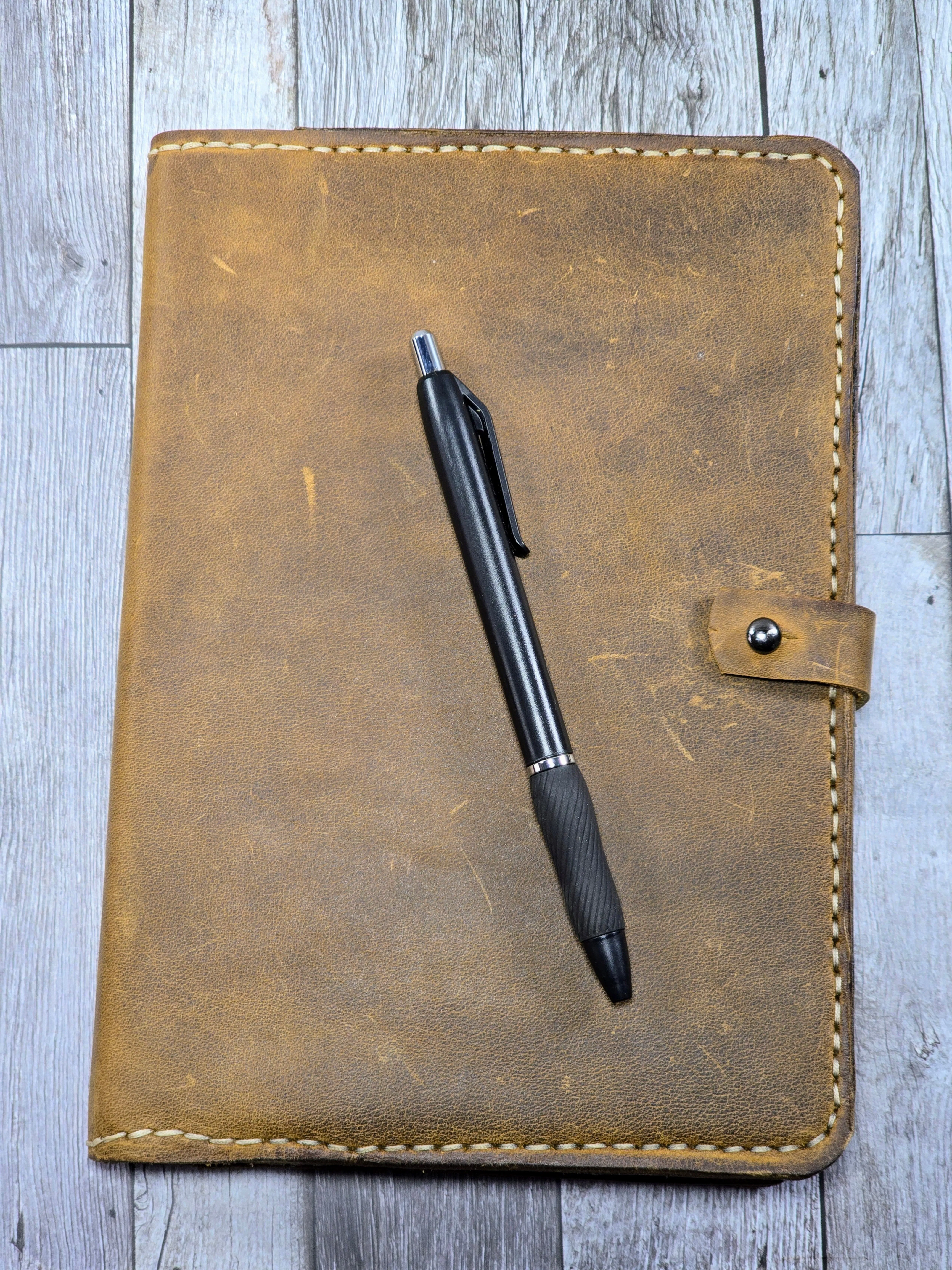 Handcrafted leather journal cover in a rich distressed tan finish, featuring visible cream stitching, a secure snap closure, and durable construction, shown with a black pen on a rustic wooden background.