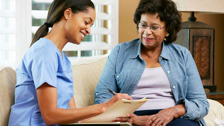 A registered nurse sits beside an elderly woman in a home environment, holding a clipboard and pen while discussing care plans