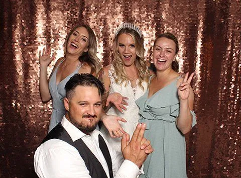 Four guests pose playfully in front of a premium rose gold sequin backdrop