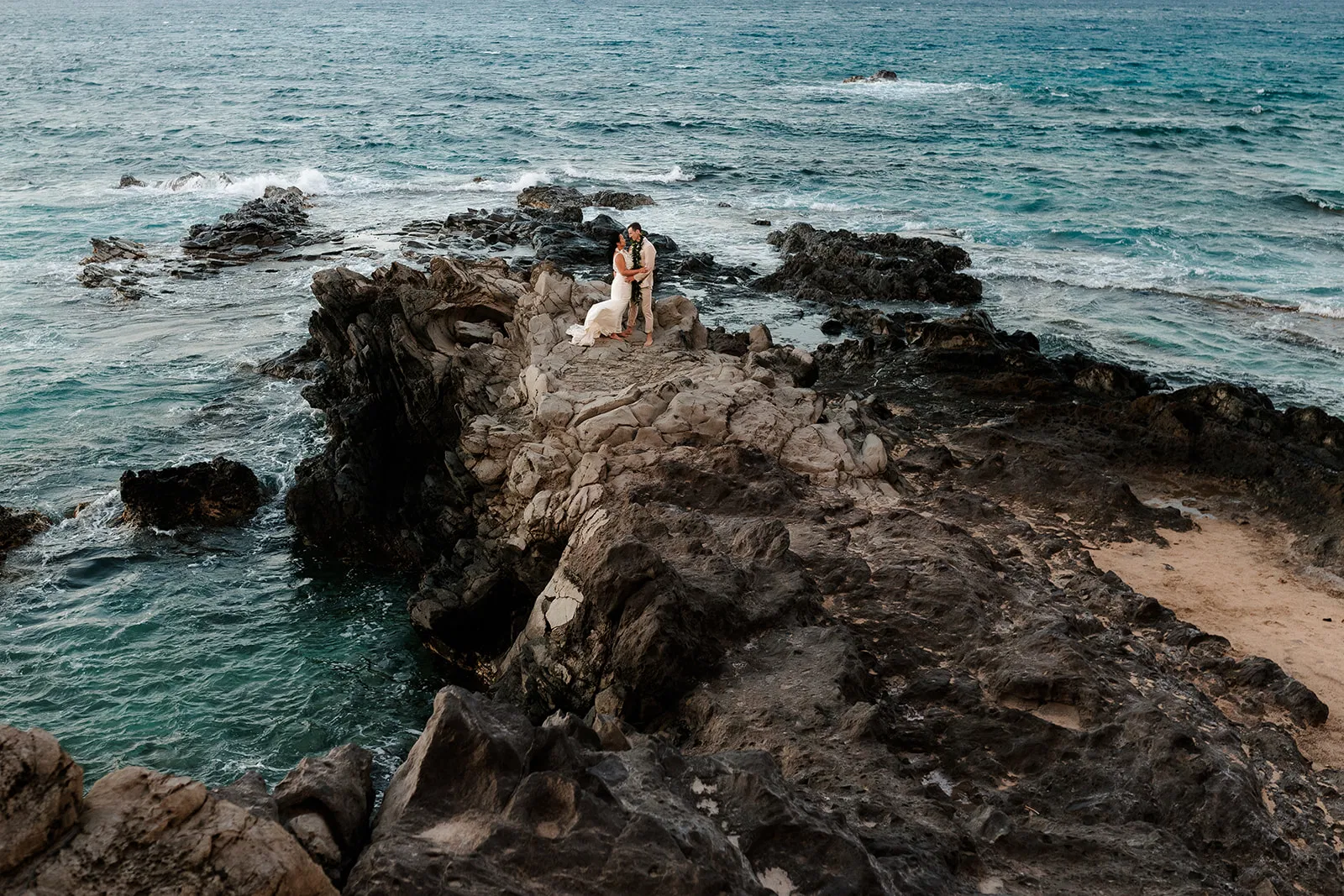 A bride and groom embrace on dramatic black lava rocks surrounded by turquoise ocean waves.