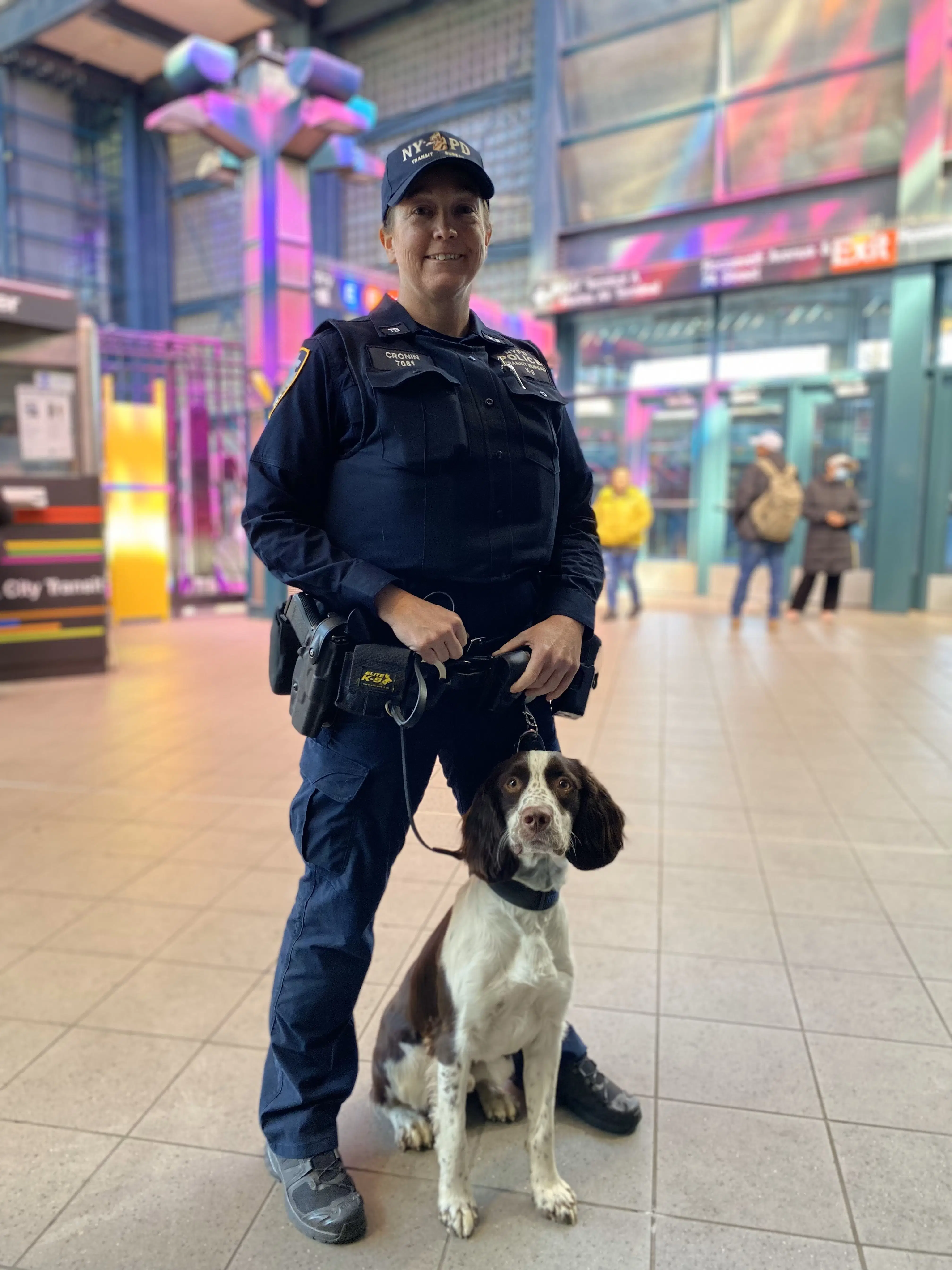 Retired police officer in NYPD tactical uniform stands with a service dog in a public transit area, demonstrating the importance of ongoing firearms safety, situational awareness, and tactical response.