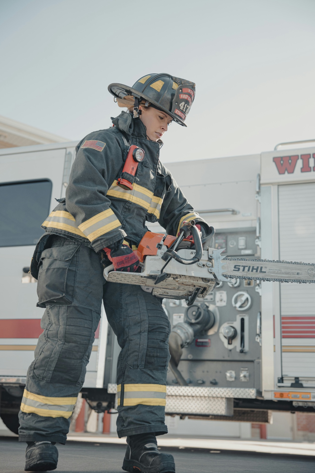Firefighter in full turnout gear operates a chainsaw in front of a fire engine