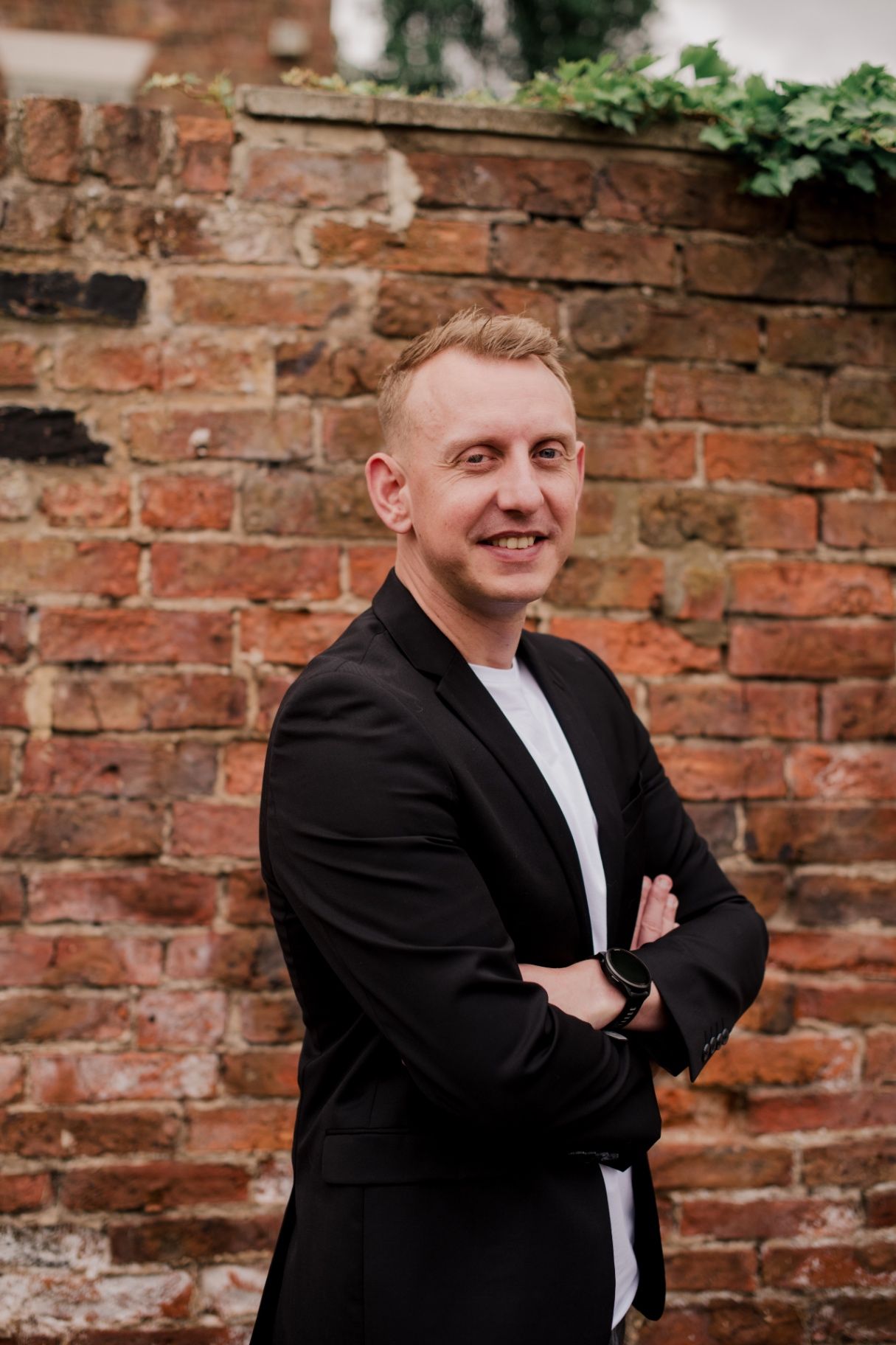 Oliver Saxon with arms crossed smiling in front of a brick wall