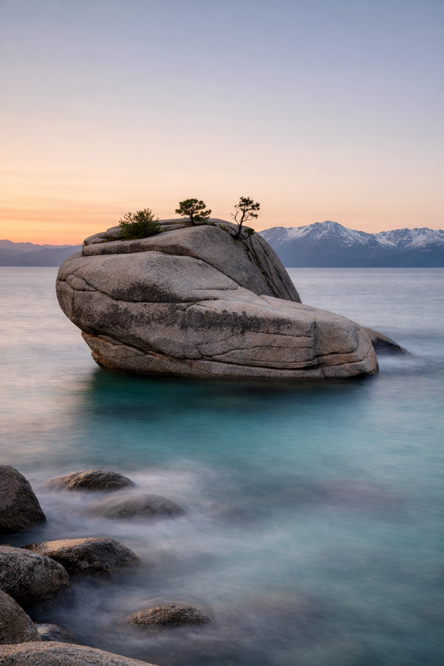 Long exposure of Bonsai Rock at sunset with misty turquoise water and Sierra Nevada mountains in Lake Tahoe.