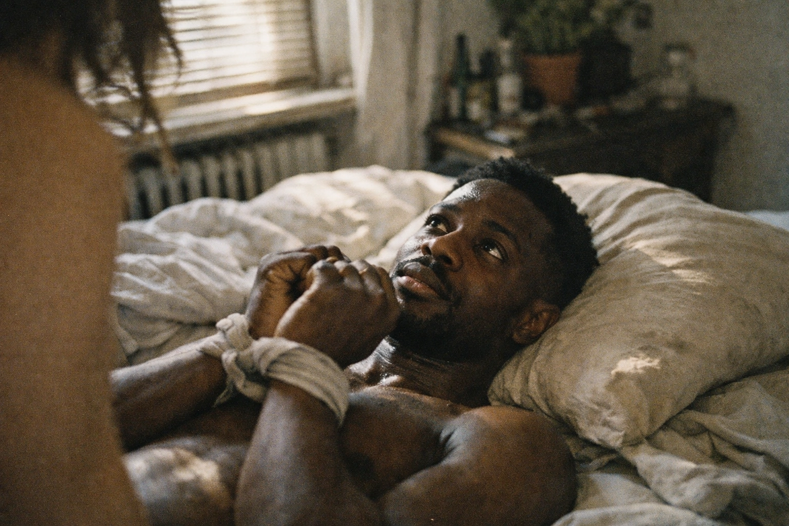 Black man in an unmade bed, wrists loosely tied with fabric, looking up with desperate tenderness, natural light, gritty documentary film look