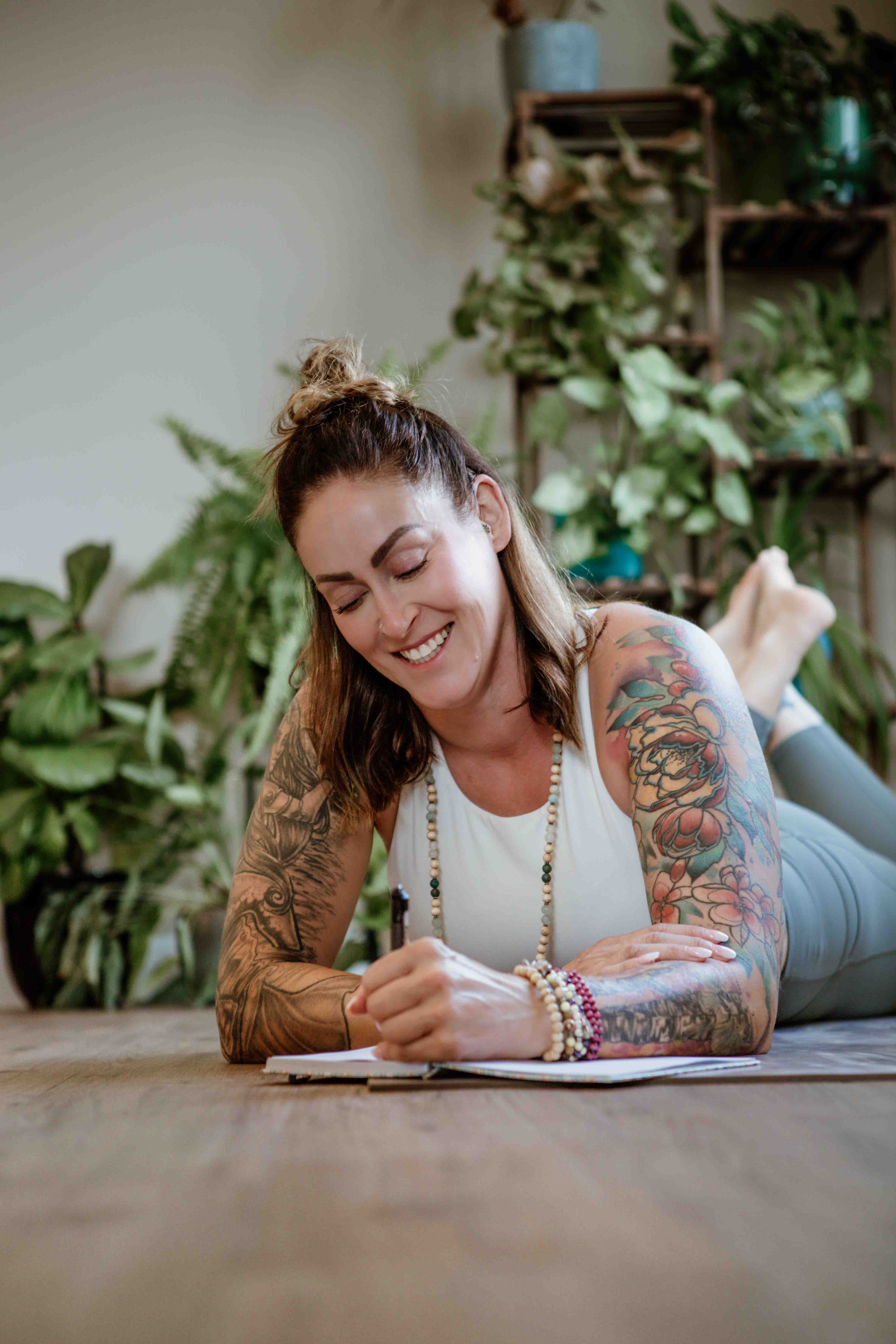 A midlife woman, dressed in comfortable activewear and adorned with mala beads and bracelets, sits on a yoga mat in a plant-filled studio. She smiles as she journals, engaging in self-reflection and considering her energy and self-care needs as part of the Capacity Check In system prior to starting yoga practice.