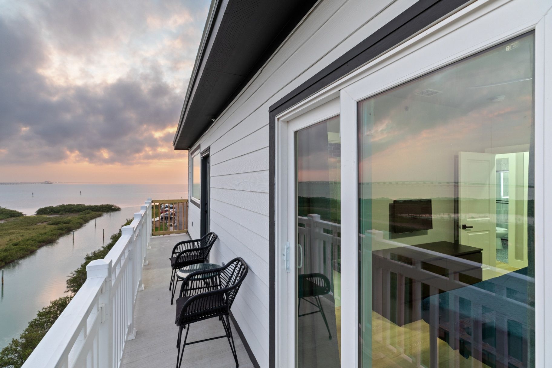 Florida Riverfront Deck View Serene balcony view at sunrise/sunset overlooking Florida riverfront/coastal greenery. Features two black chairs and a small table.