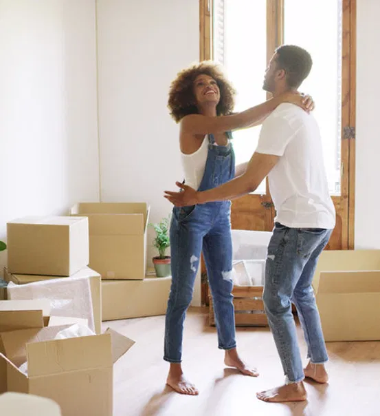 A happy couple celebrates together in a bright, unfurnished room surrounded by moving boxes.