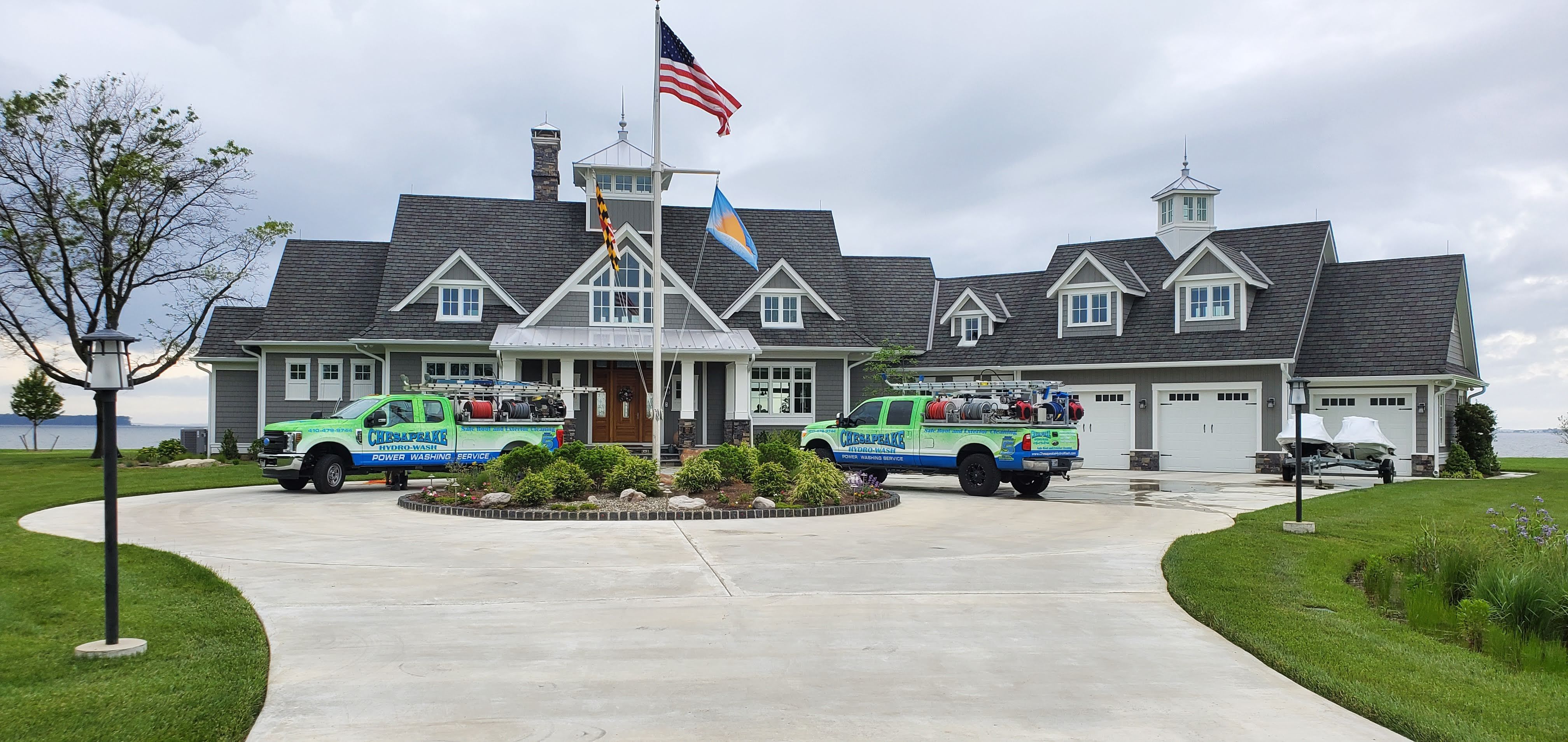 Two Chesapeake Hydro-Wash service trucks fully equipped with soft wash and power washing systems, hoses, and ladders are parked outside a residential home. The trucks display company branding and vehicle wraps promoting roof cleaning, gutter cleaning, window cleaning, and exterior washing services.