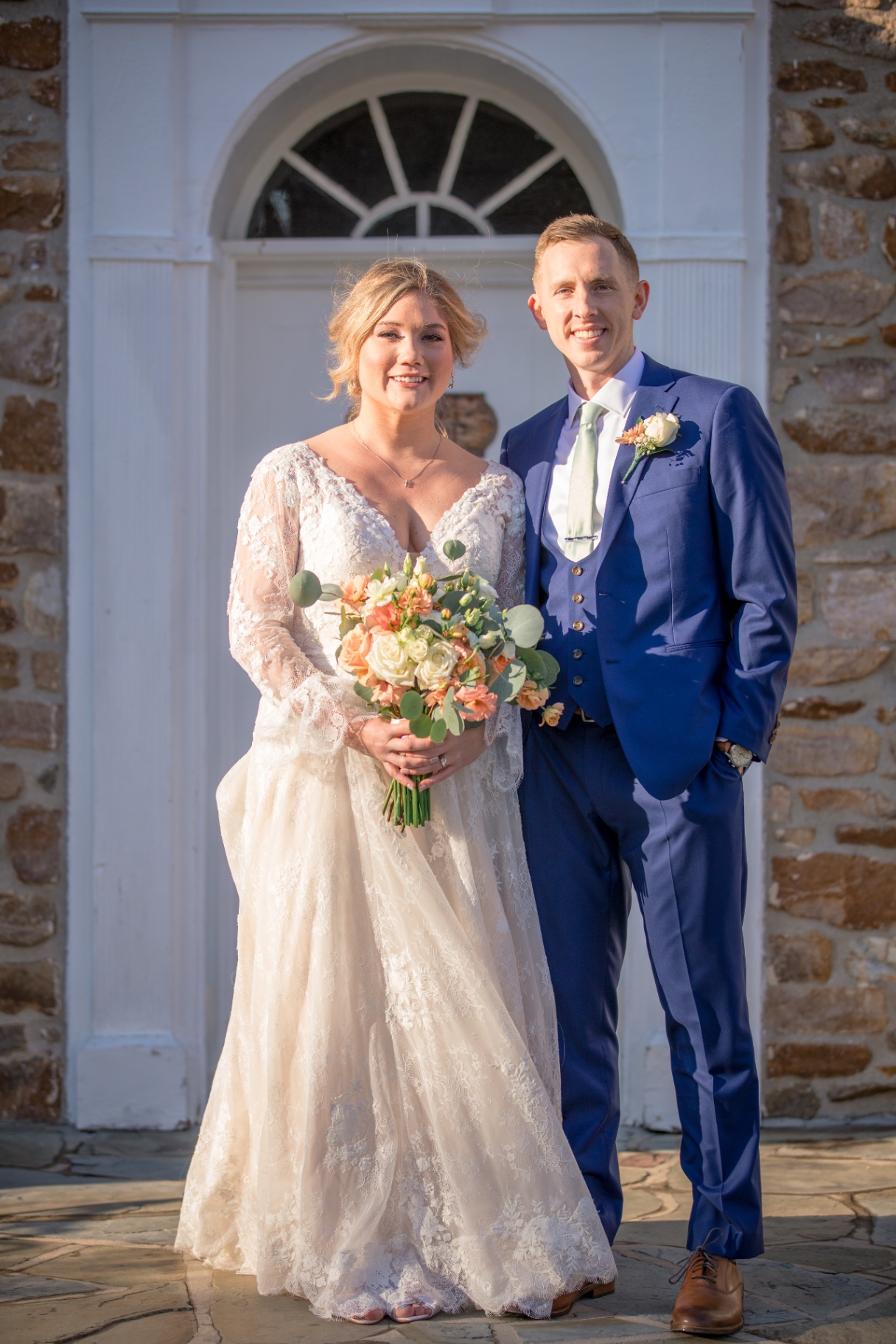 Bride and groom pose together in front of a classic stone-and-white doorway, bathed in natural sunlight
