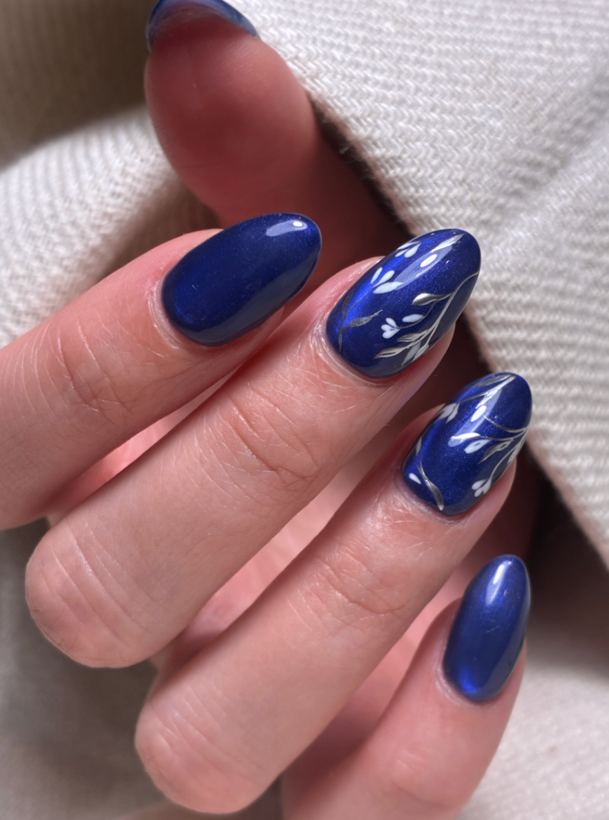 Midnight Shimmer Floral Manicure Deep shimmering blue almond-shaped manicure with delicate white and silver floral branch nail art.