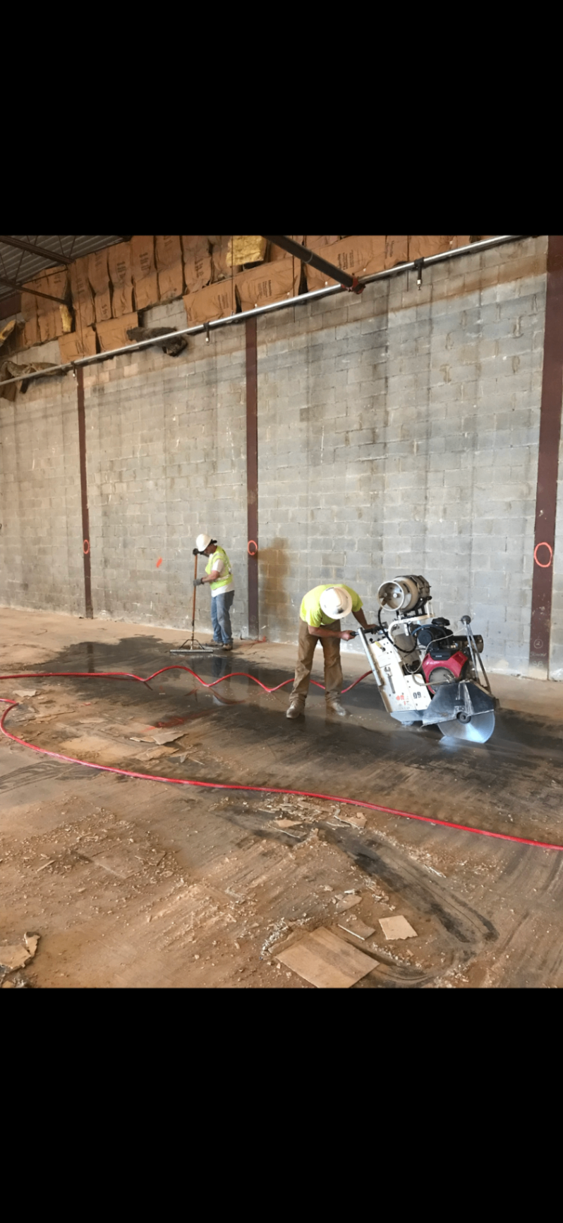 Concrete Slab Cutting Operation Two workers in safety gear are performing concrete slab cutting inside a commercial building. One operates a large walk-behind concrete saw along marked lines while the other manages cleanup and preparation. The site is clean and organized, with clear safety protocols in place.