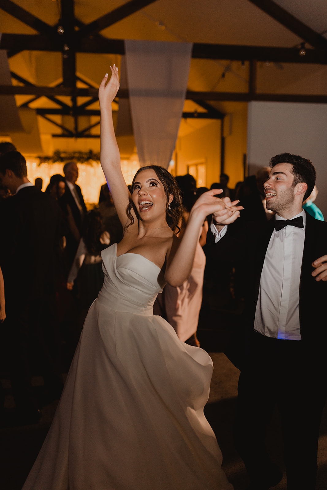 A bride and groom joyfully dance among guests on a packed dance floor A bride and groom are celebrating their wedding by dancing joyfully among their guests. The energetic atmosphere is enhanced by lively music and vibrant lighting provided by Premier DJs.