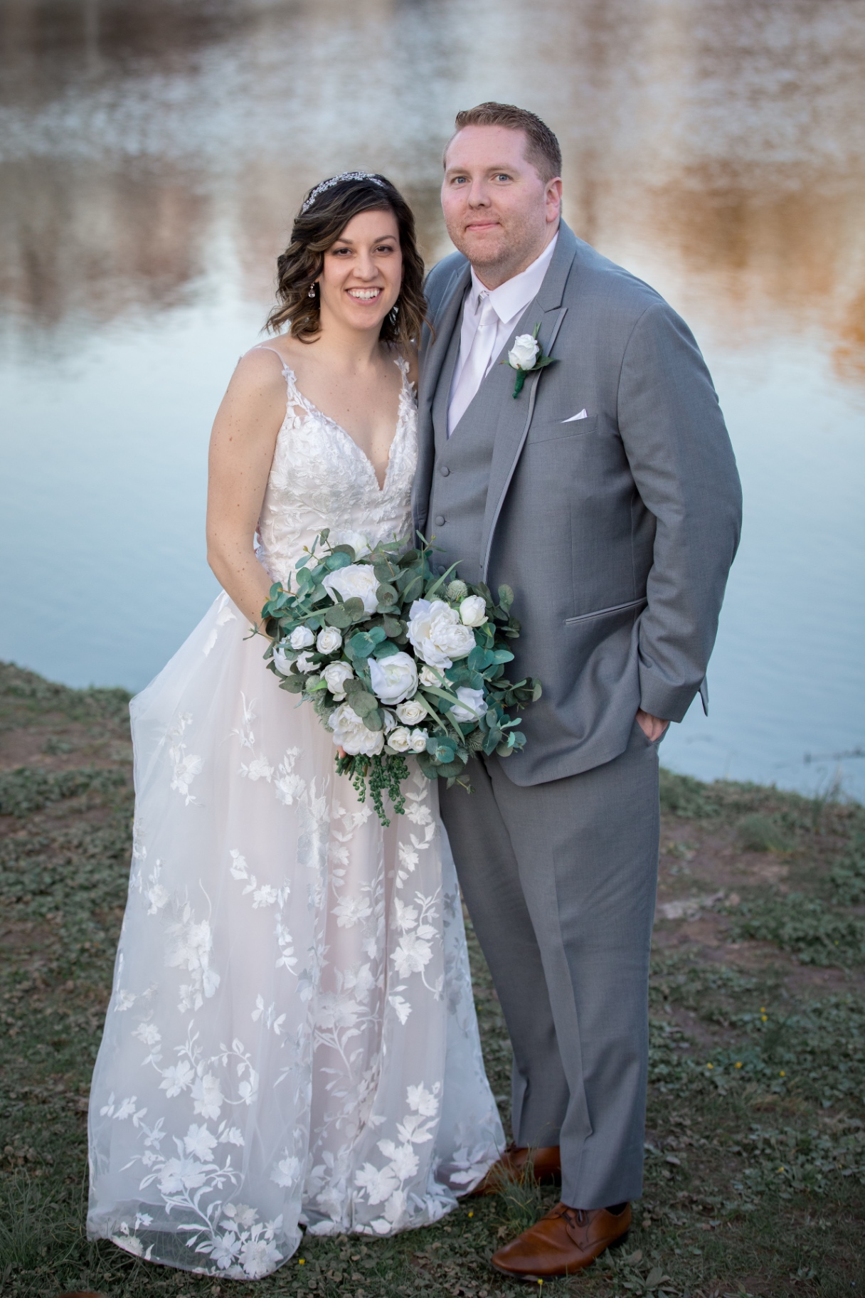 Bride and groom pose together outdoors by a calm lake, smiling