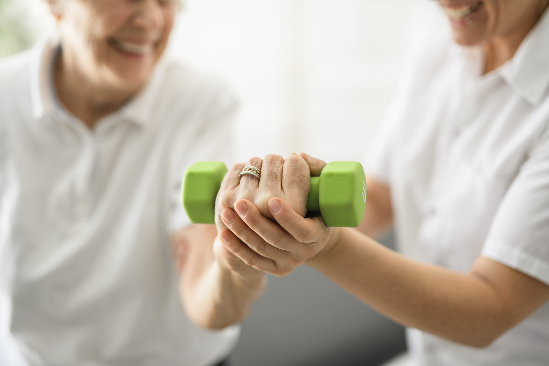 Two Trees Physical Therapy Jobs A physical therapist supports an older adult patient's hand while lifting a green dumbbell, demonstrating guided strength rehabilitation, personalized care, and encouragement in a physical therapy setting.