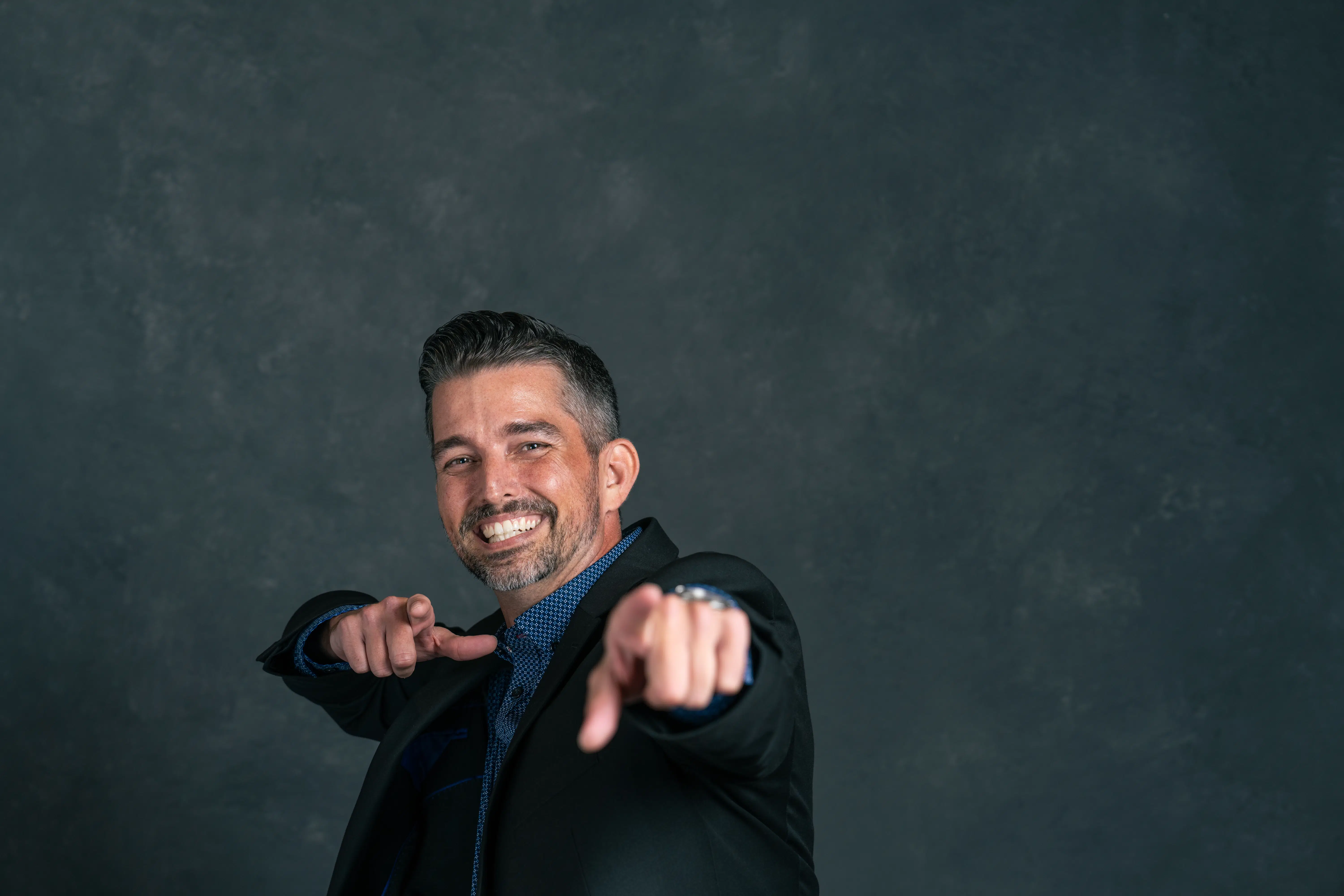 A confident man in a dark suit stands against a plain, textured background, smiling broadly and pointing directly at the camera.