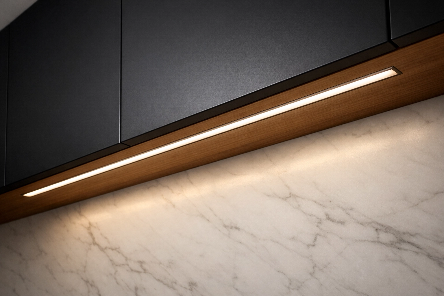 Recessed aluminum LED profile under black cabinets creating uniform warm light on a white marble backsplash.