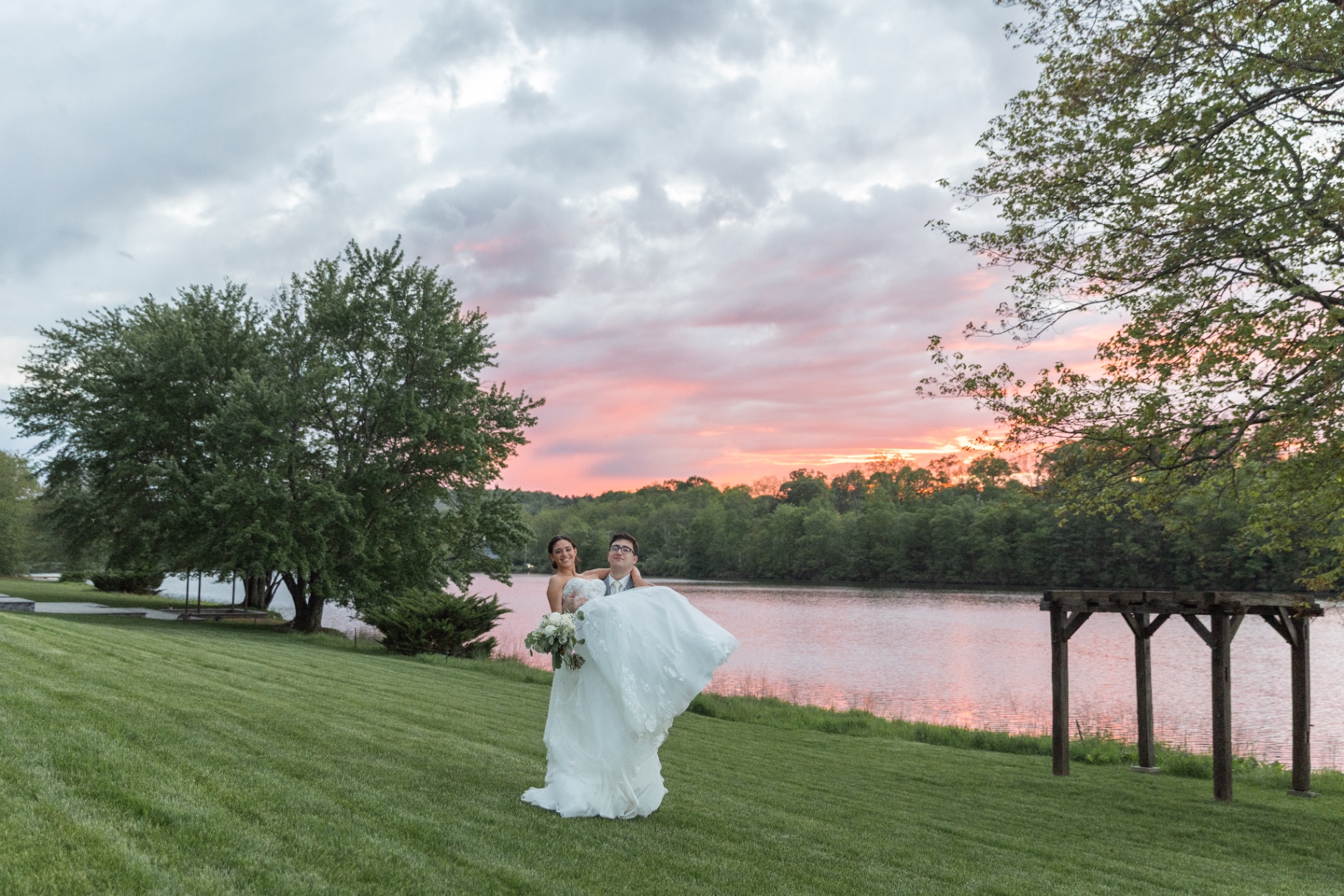 A groom carries his bride near a lakeside at sunset