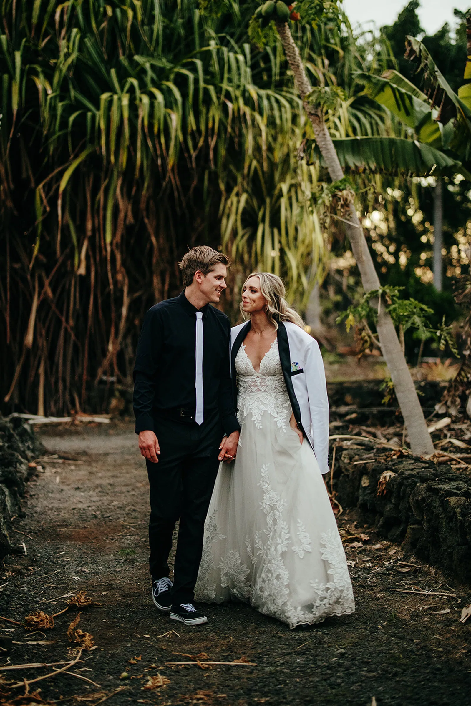 A newlywed couple strolls hand in hand along a rustic lava rock path, surrounded by lush tropical greenery.