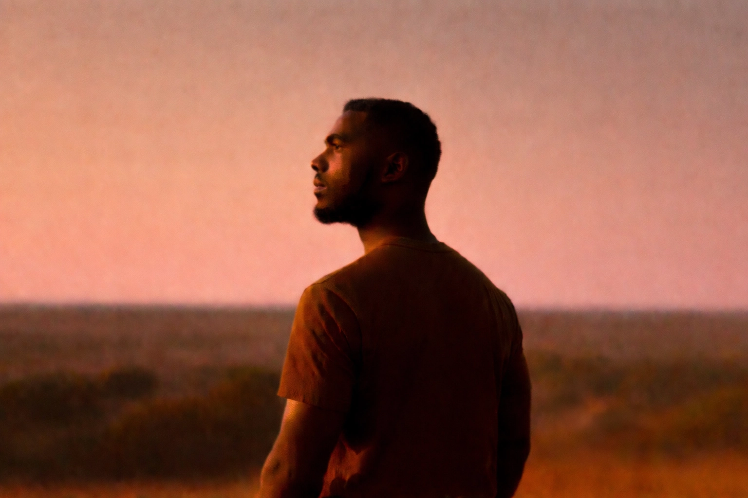 The Invisible Burden A Black man stands outside at sunset, looking thoughtful.