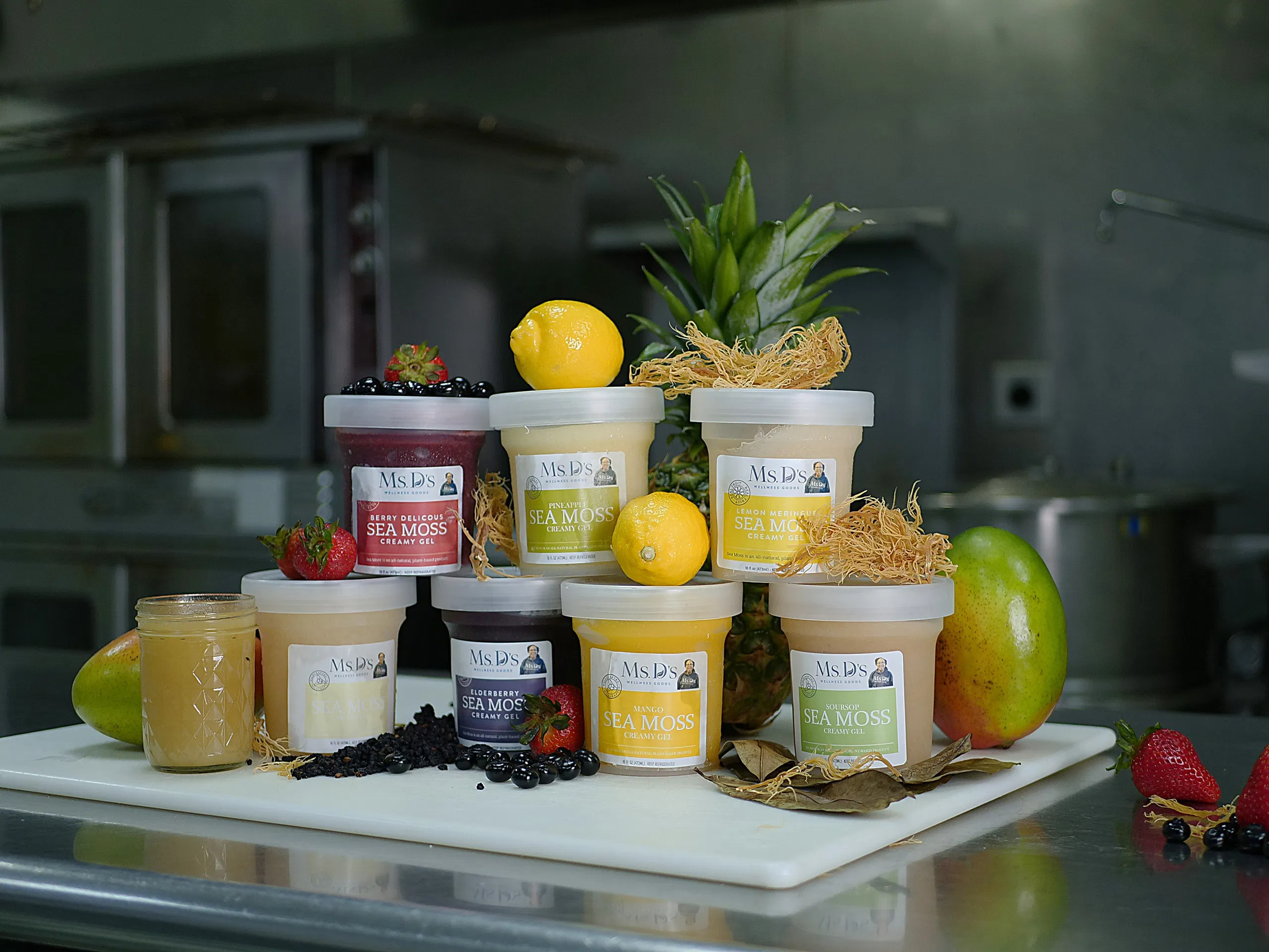 A selection of Ms. D’s Wellness Goods 16oz Sea Moss Gels in various flavors, Berry Delicious, Elderberry, Lemon Meringue, Pineapple, Mango, and Plain, artfully arranged with fresh fruits.